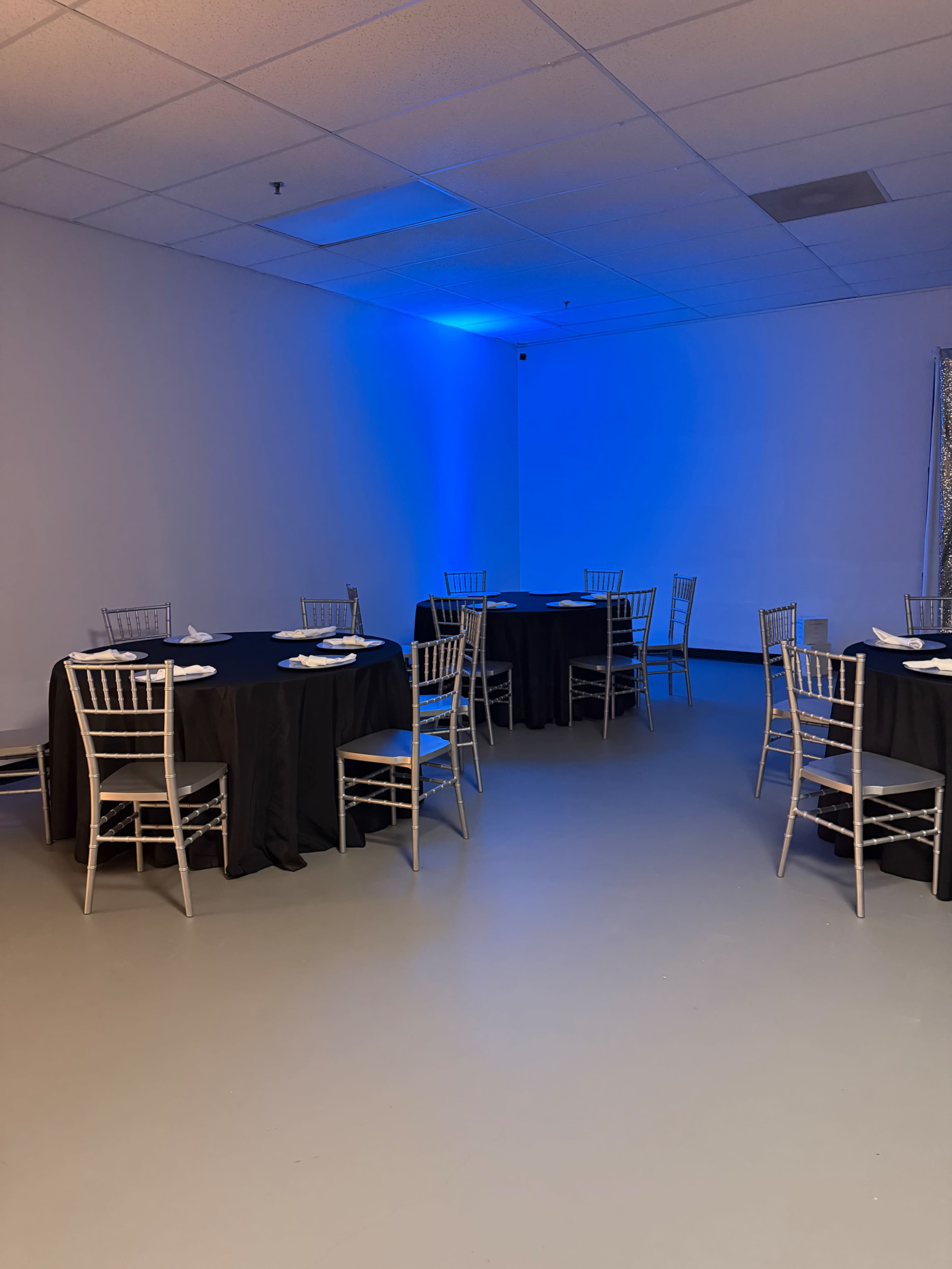A well-lit event space features several round tables covered with black tablecloths and surrounded by silver chairs.