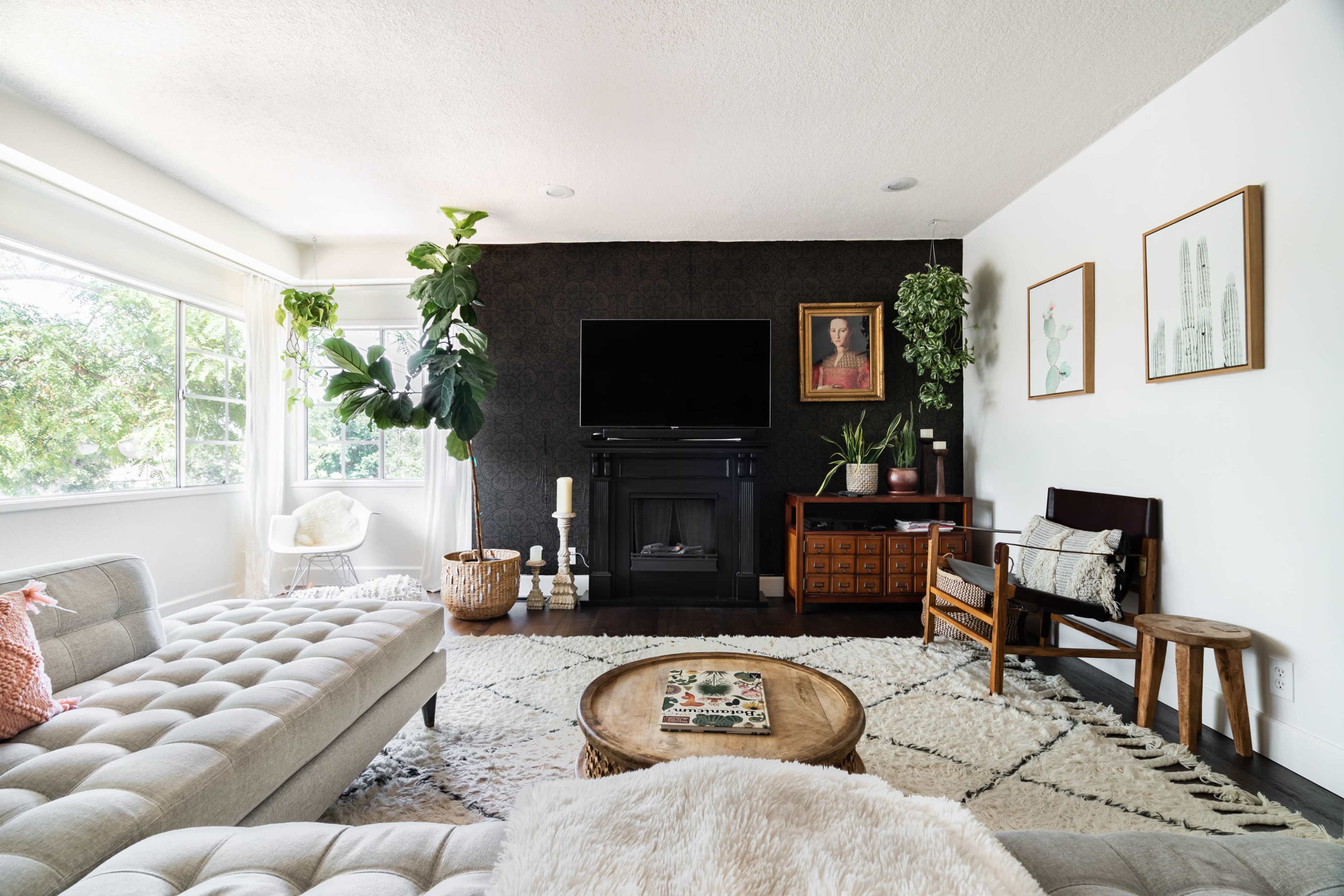 The living room features a light-colored sectional sofa, a round wooden coffee table, a fireplace, and various plants, with a large television mounted on the wall.