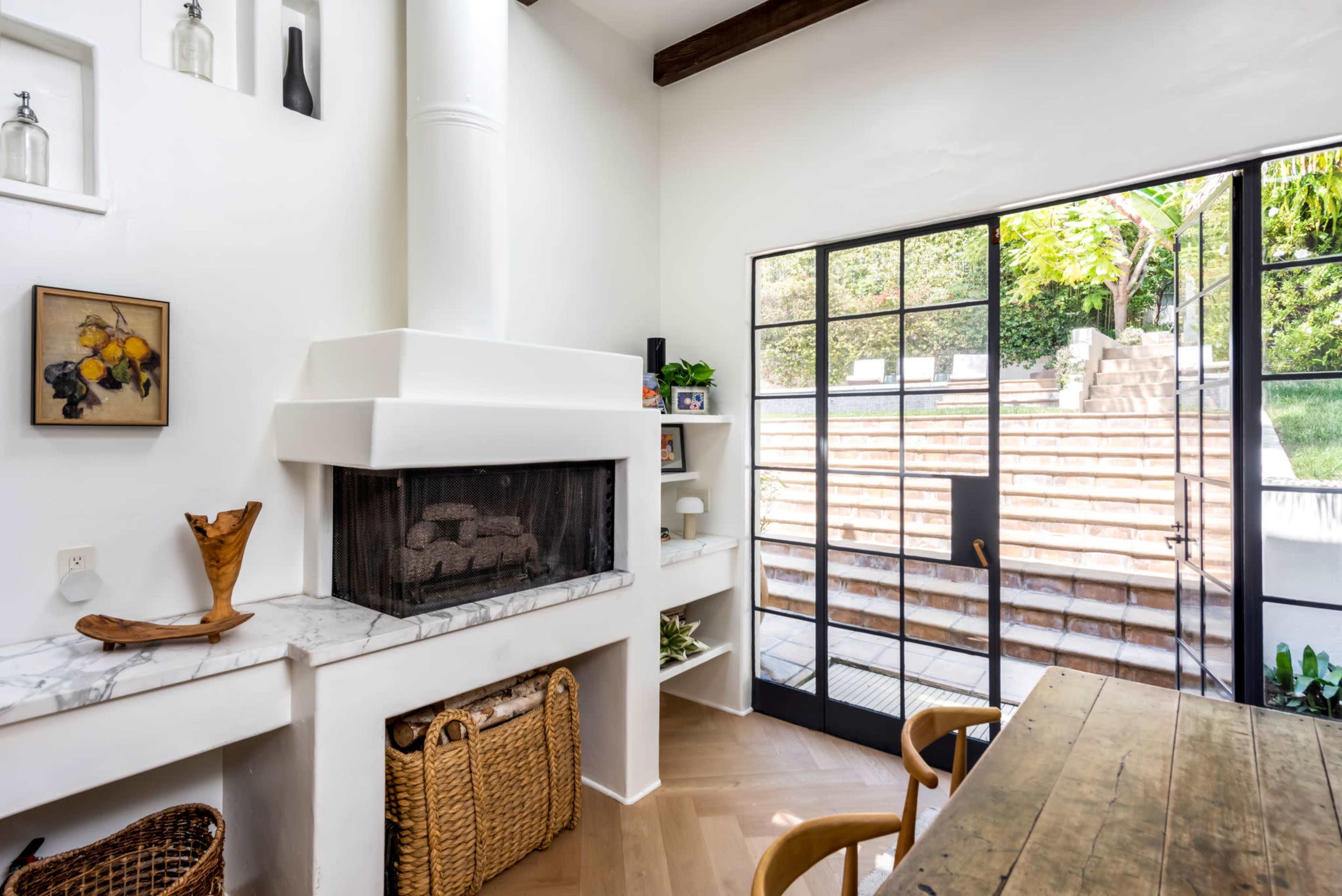 The image shows a bright interior space with a modern fireplace, wooden table, and large glass doors leading to an outdoor area with steps.