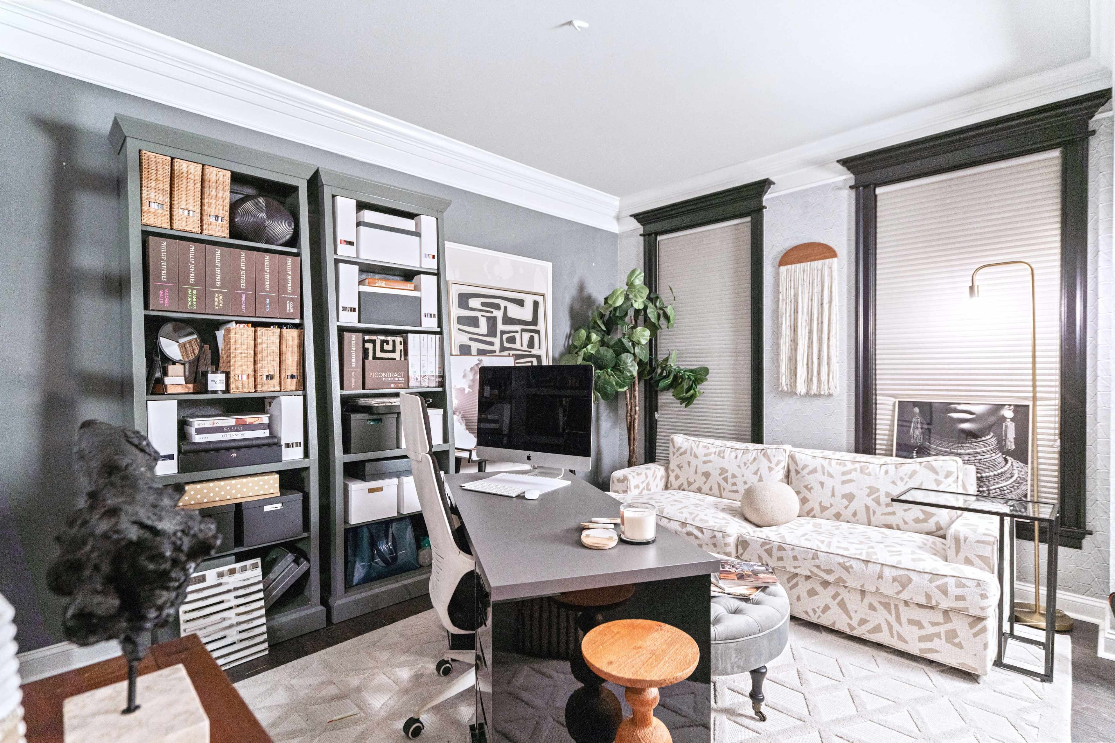 A modern office space featuring a desk with a computer, a bookshelf filled with storage boxes and books, a sofa, and large windows with blinds.