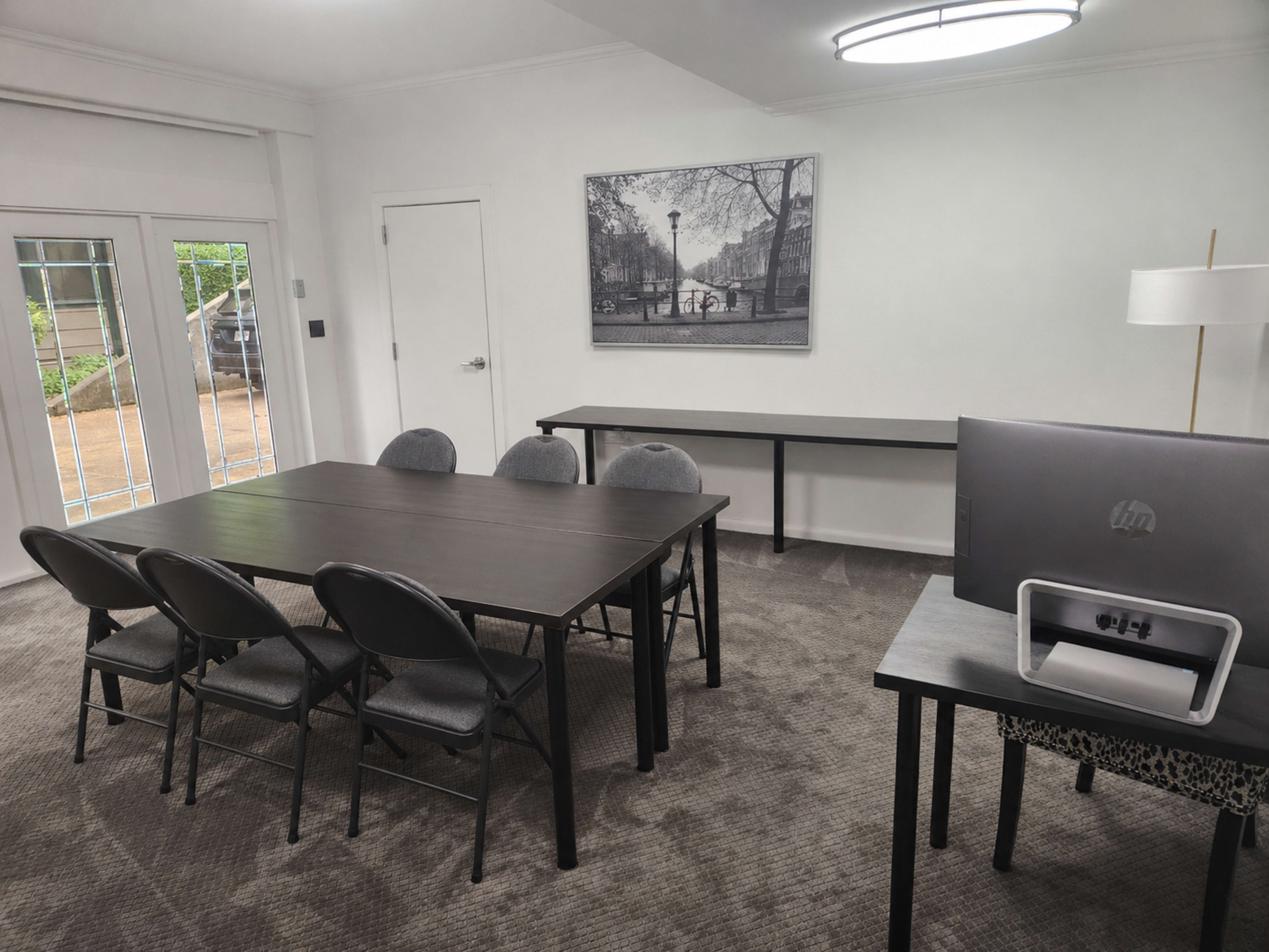 A conference room features a rectangular table surrounded by six chairs, with a laptop on a side desk and a large framed photo on the wall.
