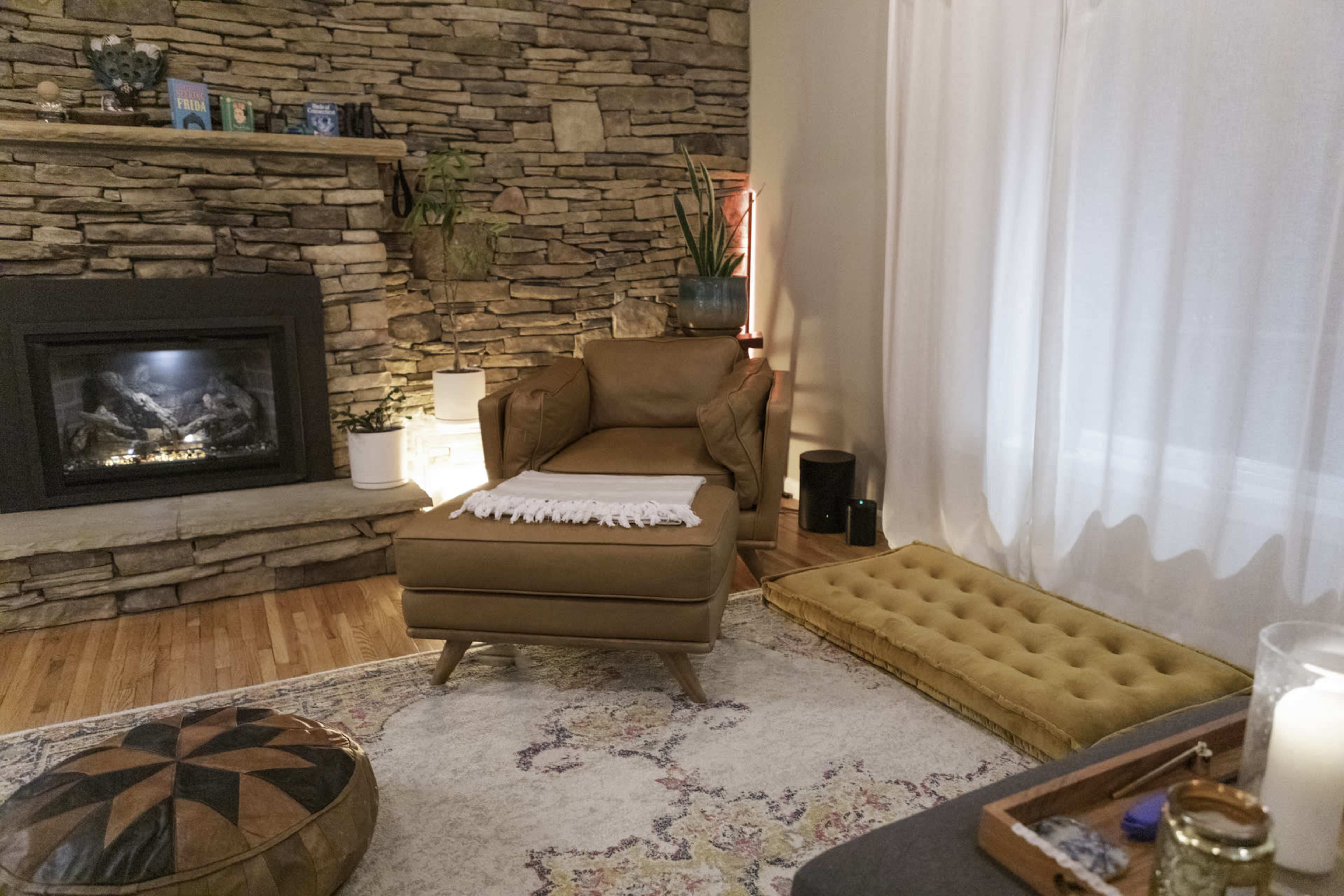 A cozy living room features a stone fireplace, a brown armchair with a white throw, and a decorative rug on the wooden floor.