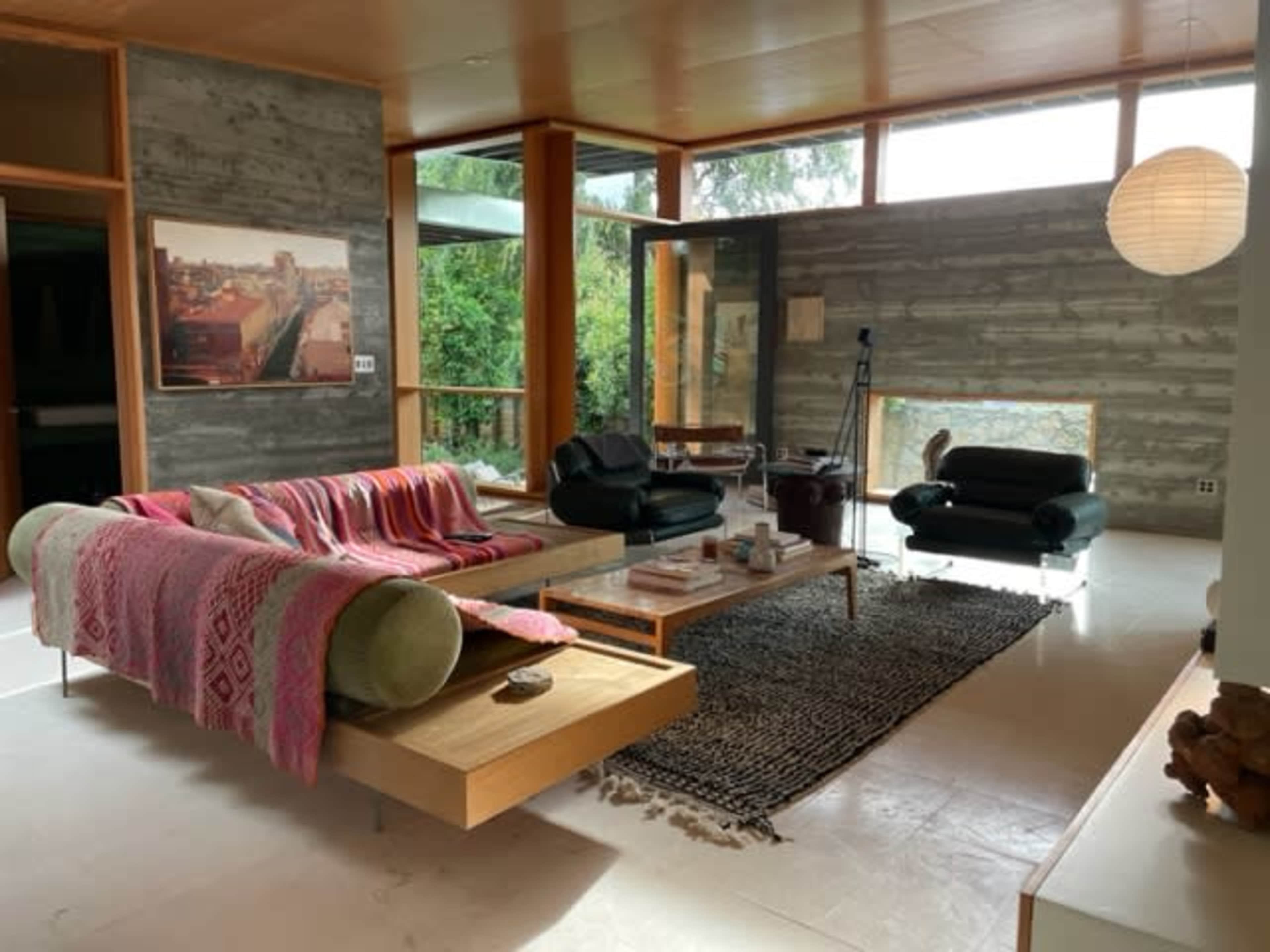 The living room features a wooden floor, a large window allowing natural light, a sectional sofa with a vibrant blanket, and a coffee table surrounded by a textured rug.