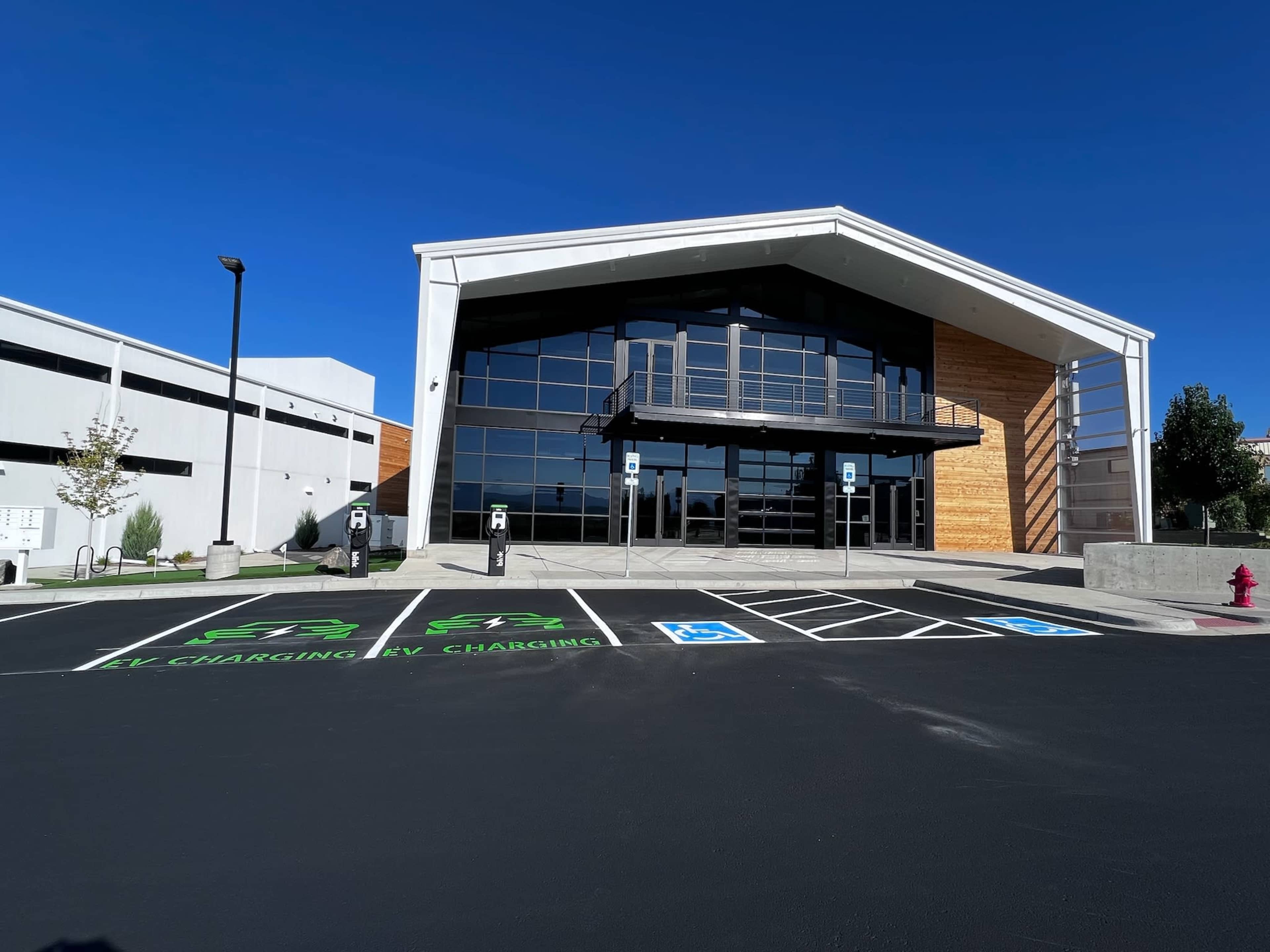A modern building with large glass windows and a wooden facade, surrounded by a parking lot with designated electric vehicle charging stations and accessible parking spaces.