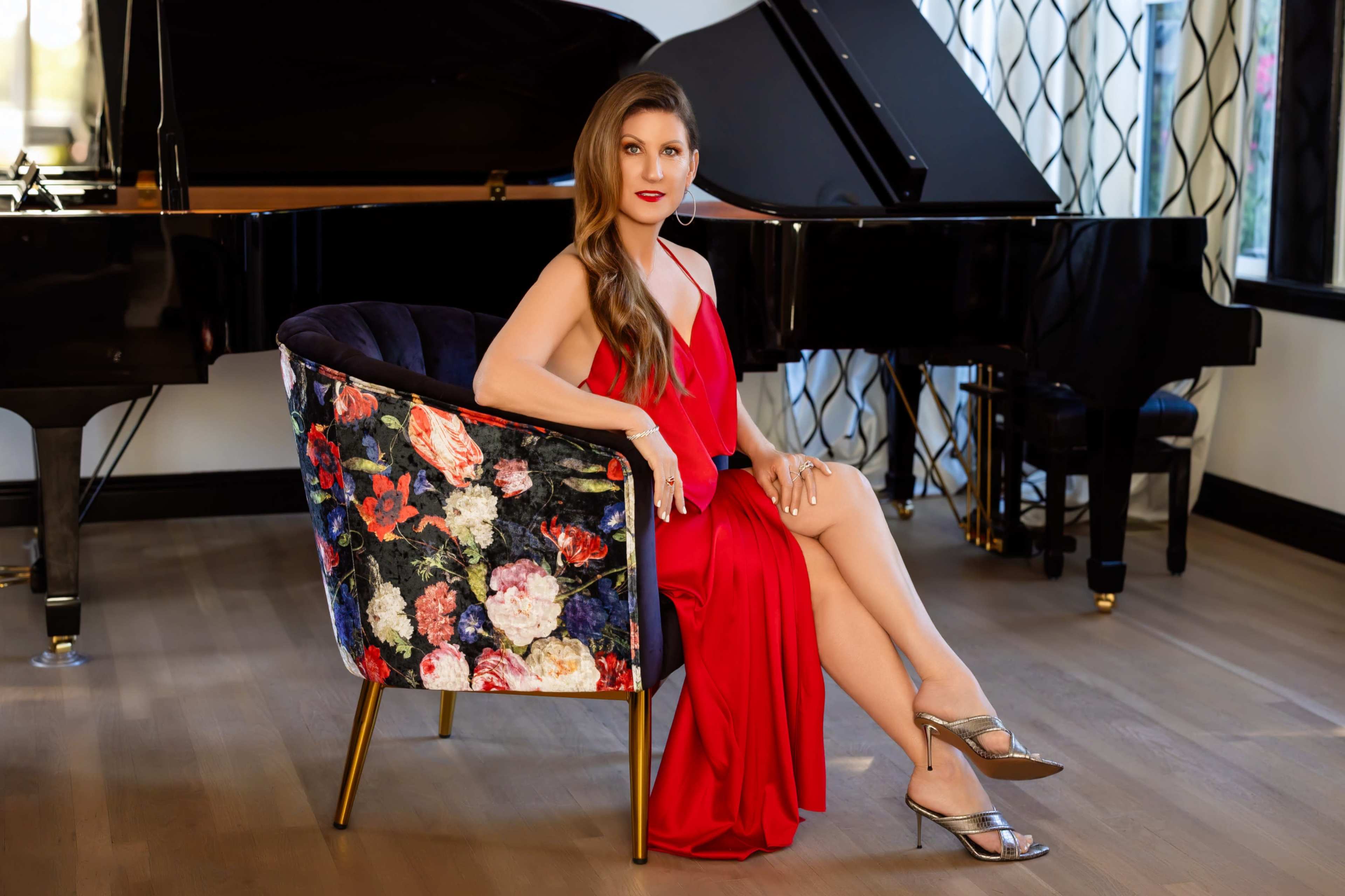 A woman in a red dress sits elegantly in a floral armchair beside a grand piano.