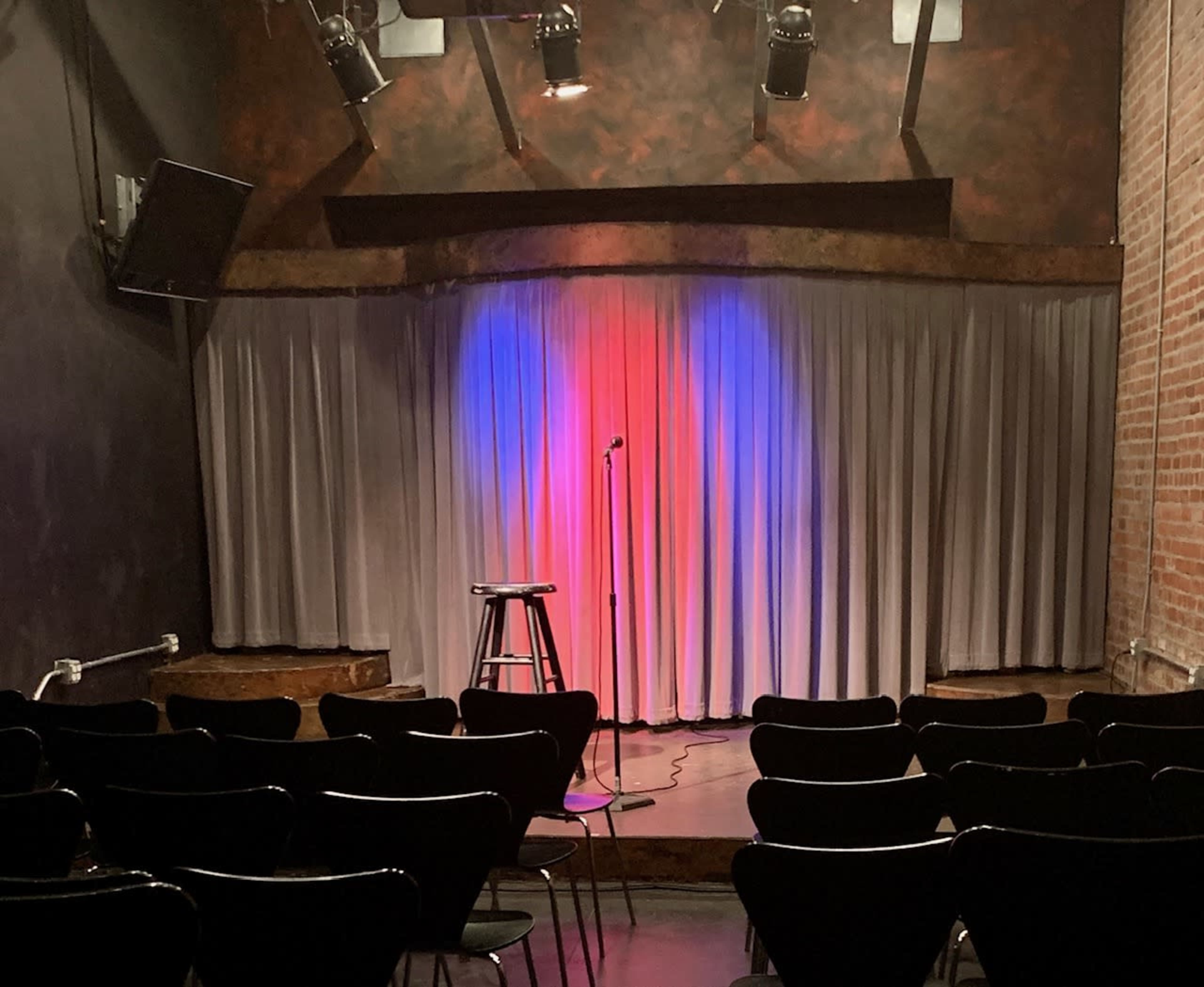 A small comedy stage with a microphone stand and a stool is set against a backdrop of red and blue lights illuminating the curtain.
