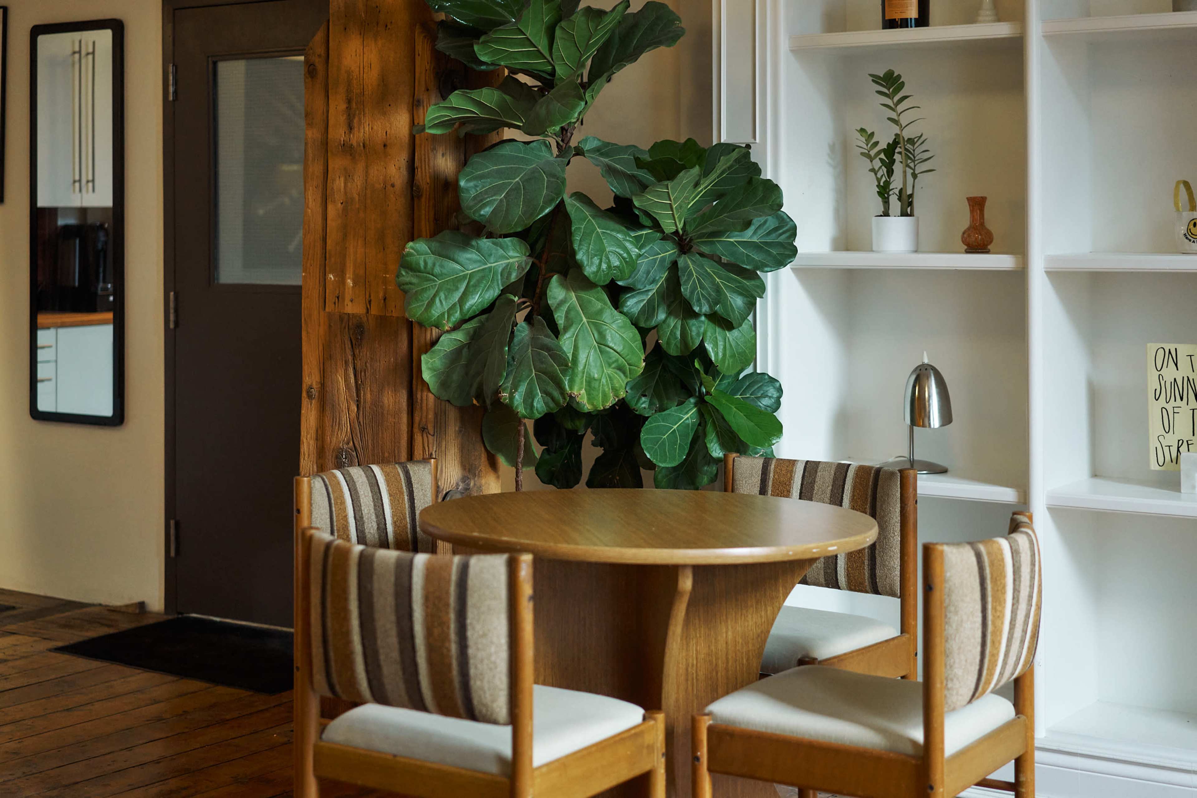 A round wooden table is surrounded by striped chairs in a room with a large potted plant and built-in shelving.