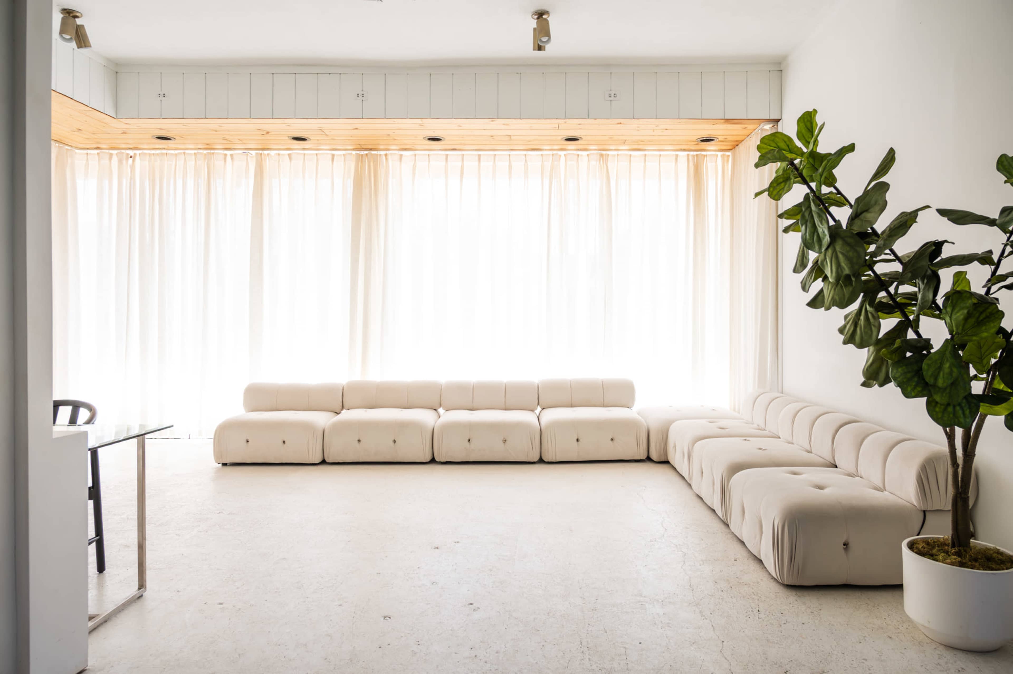 A bright, minimalist lounge area features a sectional sofa with tufted cushions and large windows draped with sheer curtains.