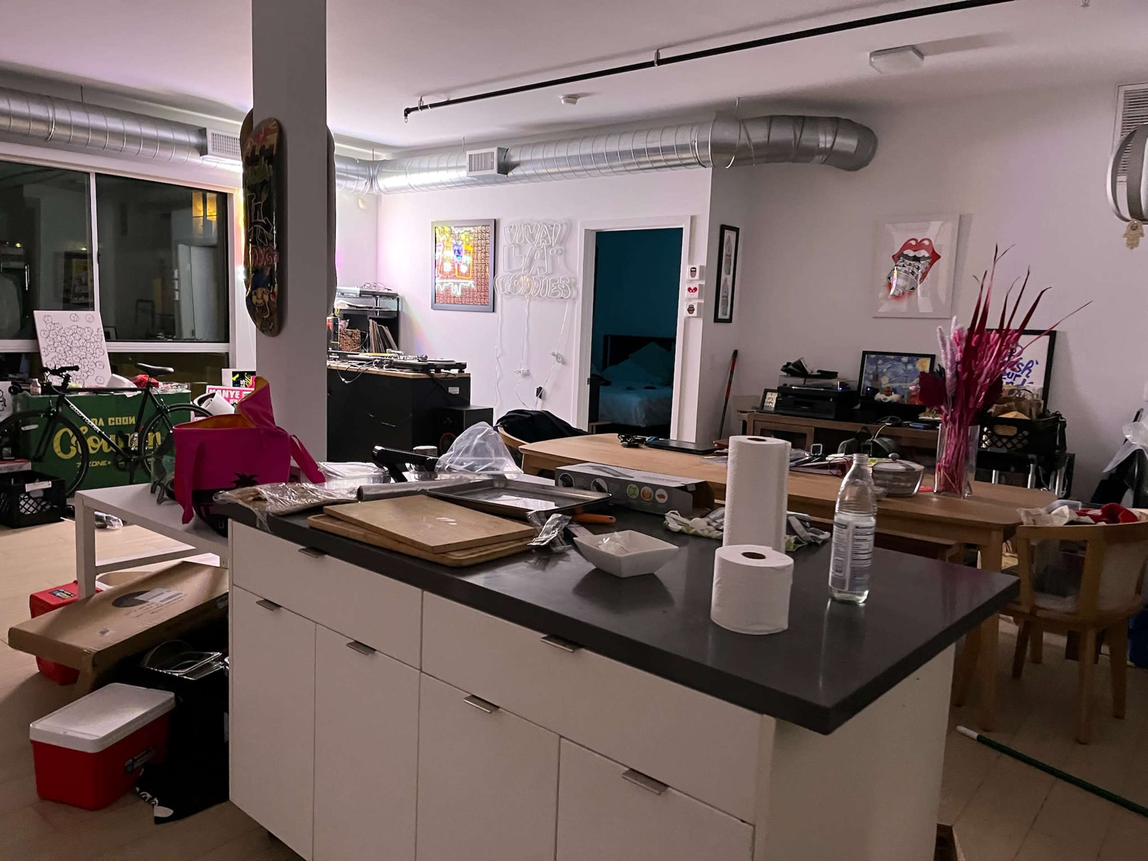 The image shows a cluttered kitchen and living area with various items and furniture scattered around, including a countertop, a dining table, and a large window.