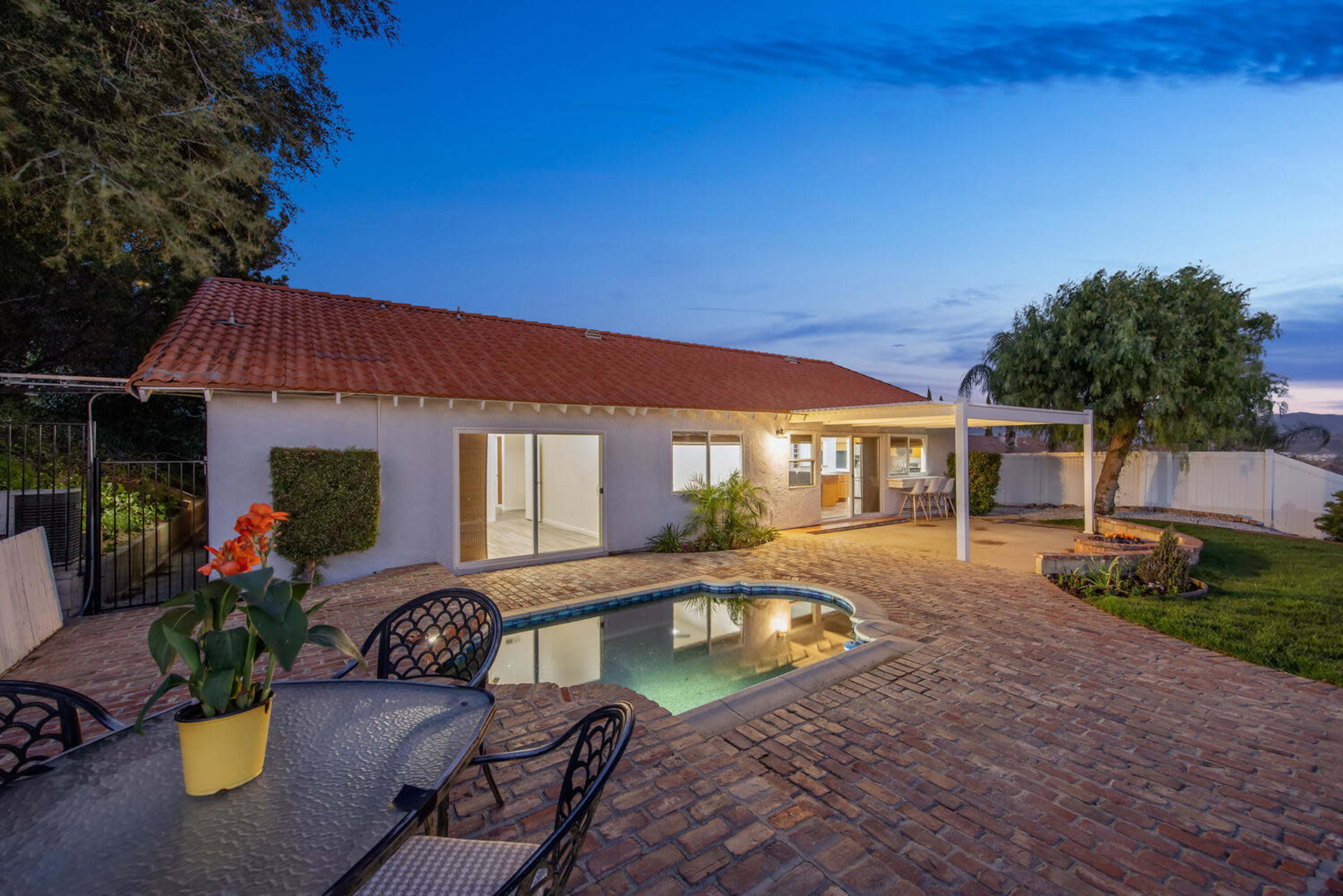 A house with a red-tiled roof overlooks a swimming pool and patio area, set against a twilight sky.