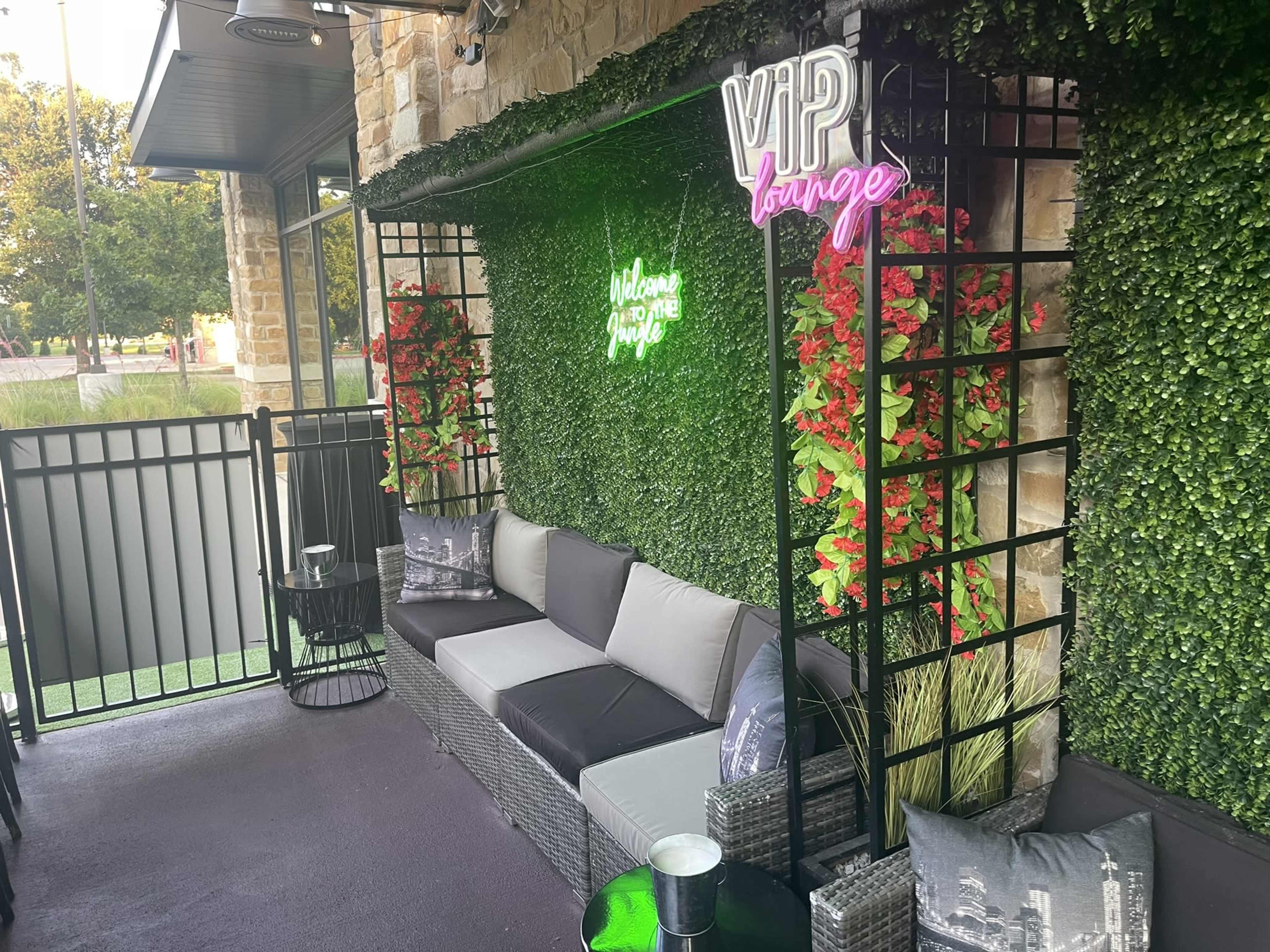 A cozy outdoor seating area with green wall panels, neon signs, and decorative plants.