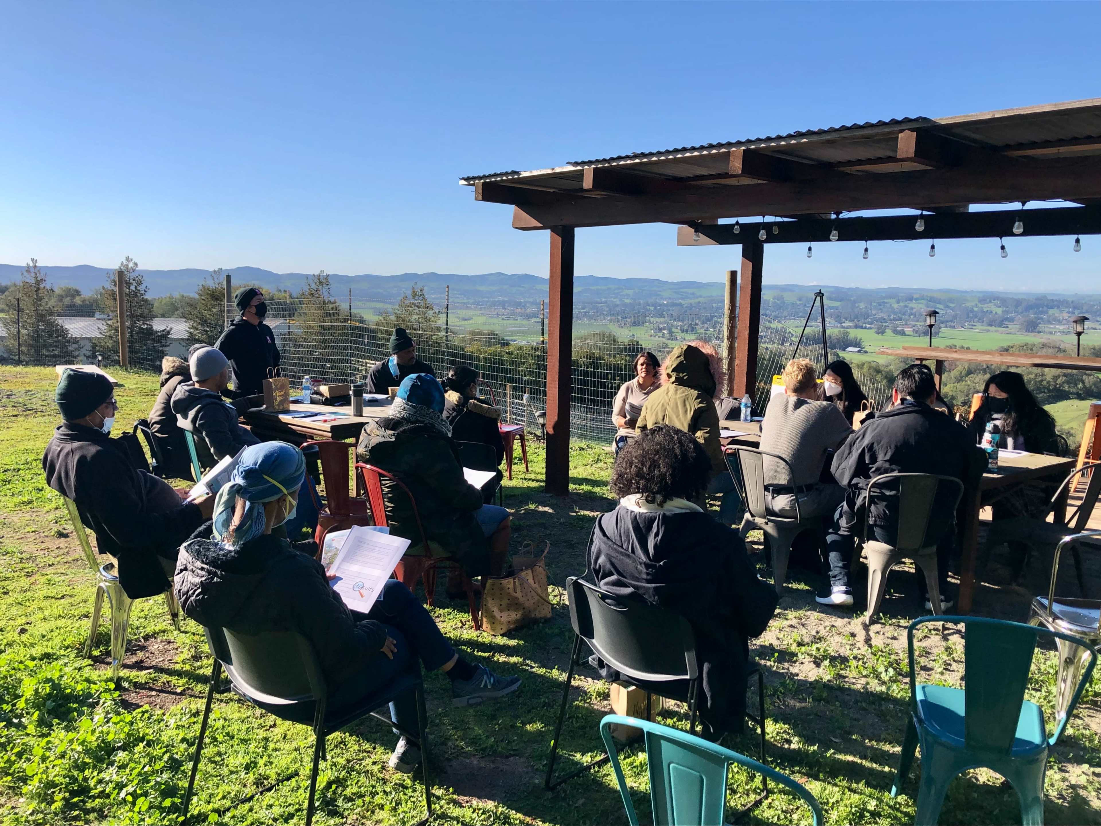 A group of people sits at tables under a shelter, facing a speaker with hills and fields in the background.