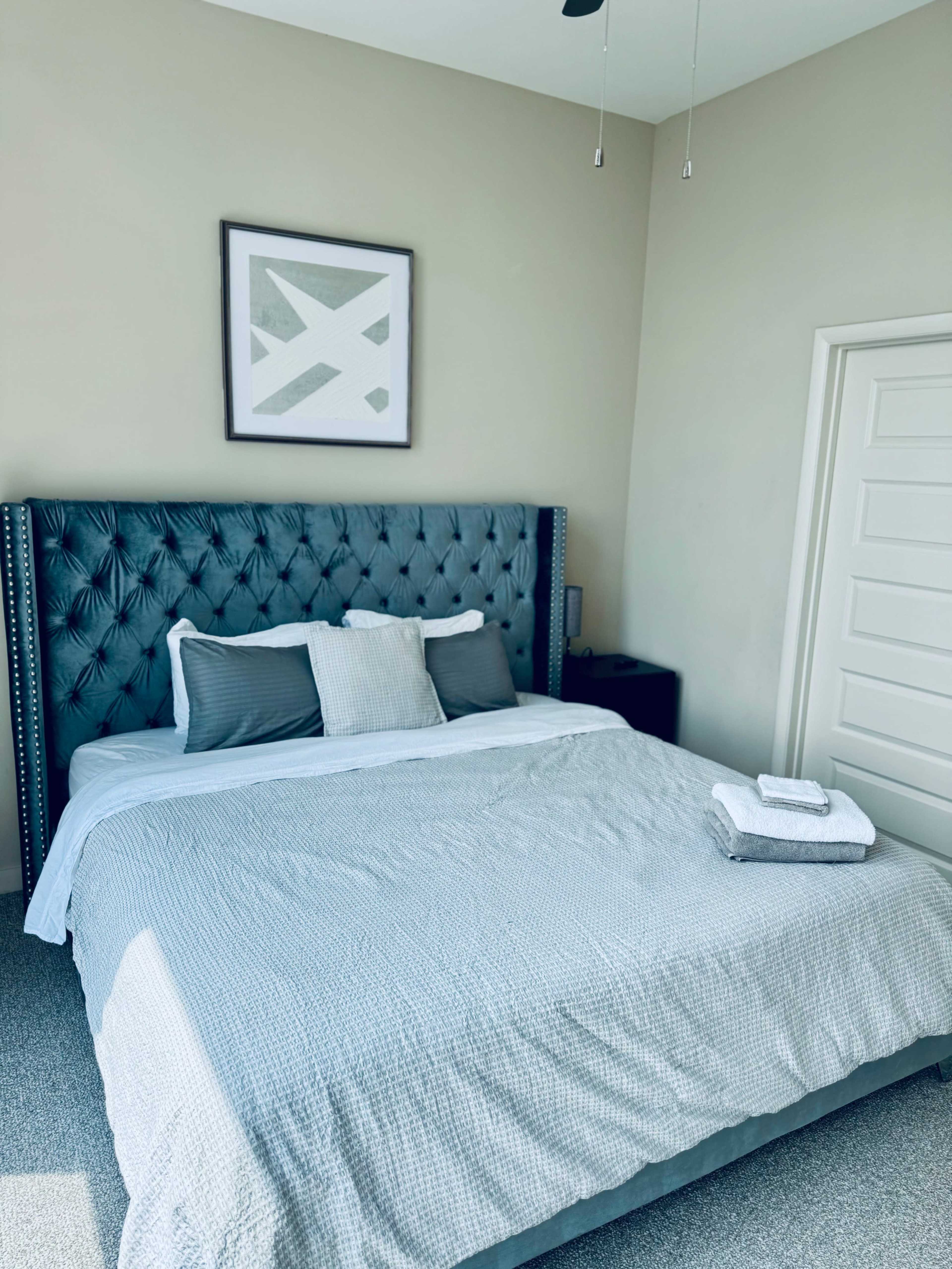 A neatly made bed with a tufted blue headboard and gray bedding is positioned in a well-lit bedroom featuring minimalist decor and a closed door.