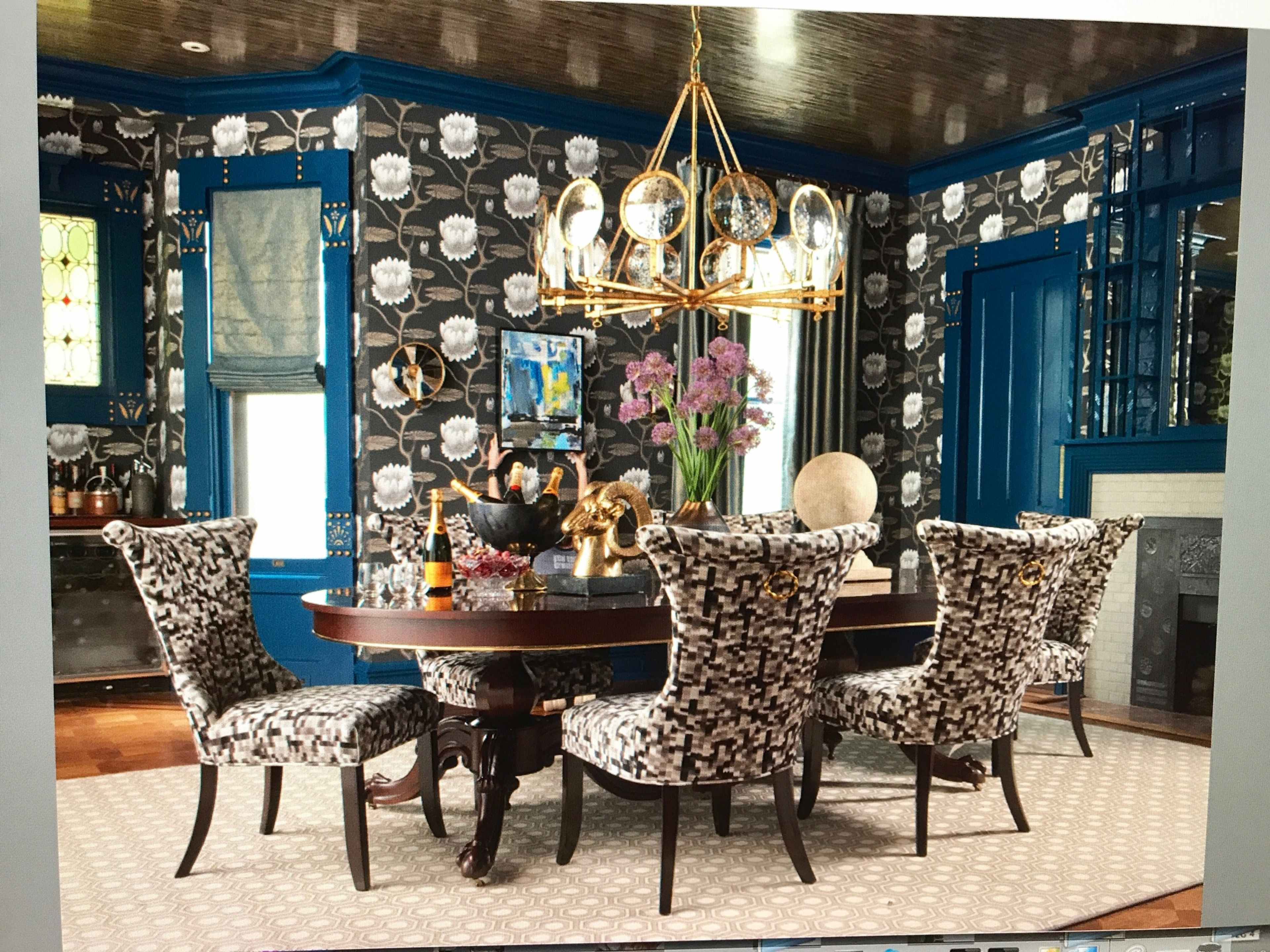 A dining room with a round table surrounded by patterned chairs, a chandelier overhead, and floral wallpaper.