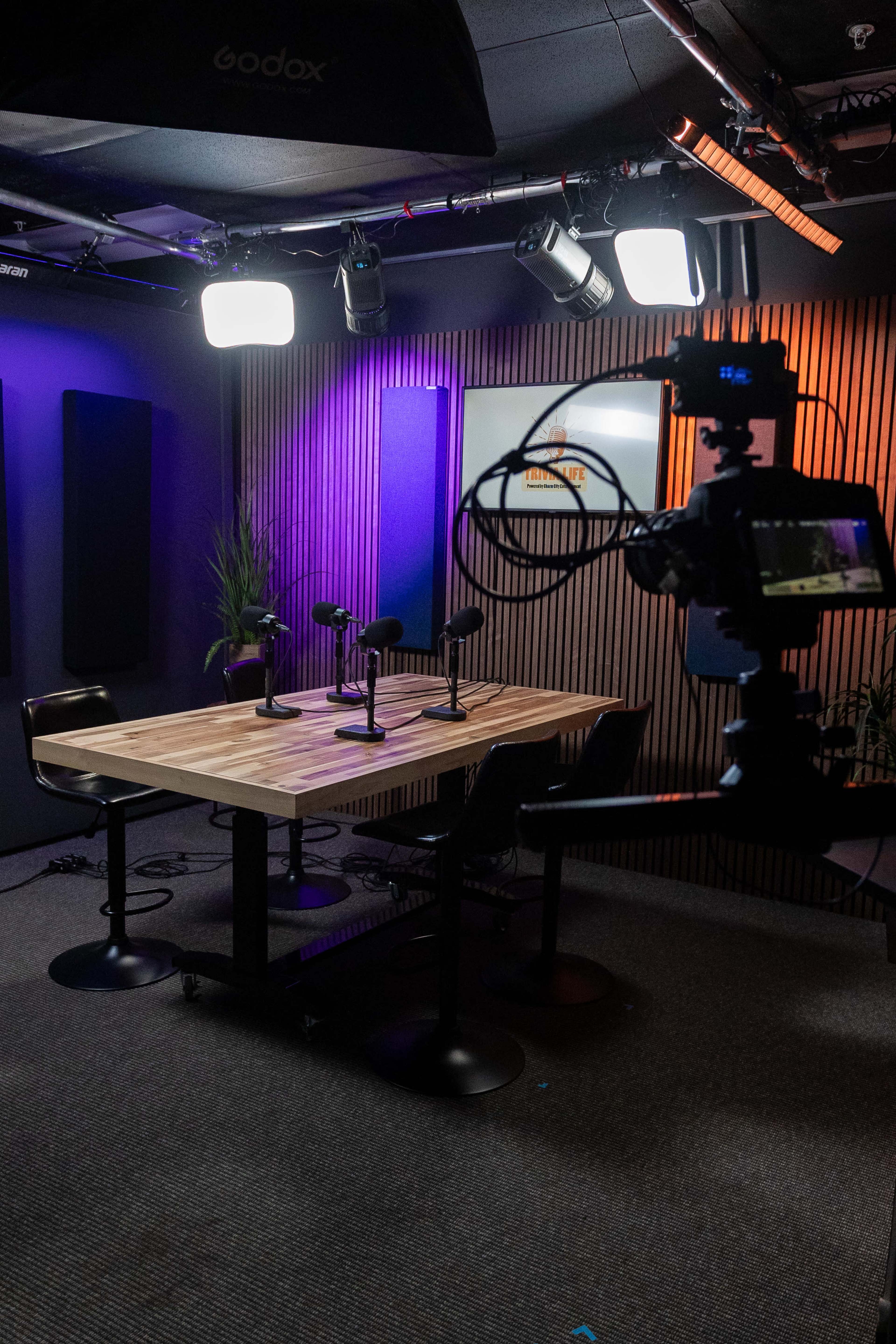 A modern recording studio features a wooden table with four microphones, surrounded by black chairs and equipped with various lighting fixtures.
