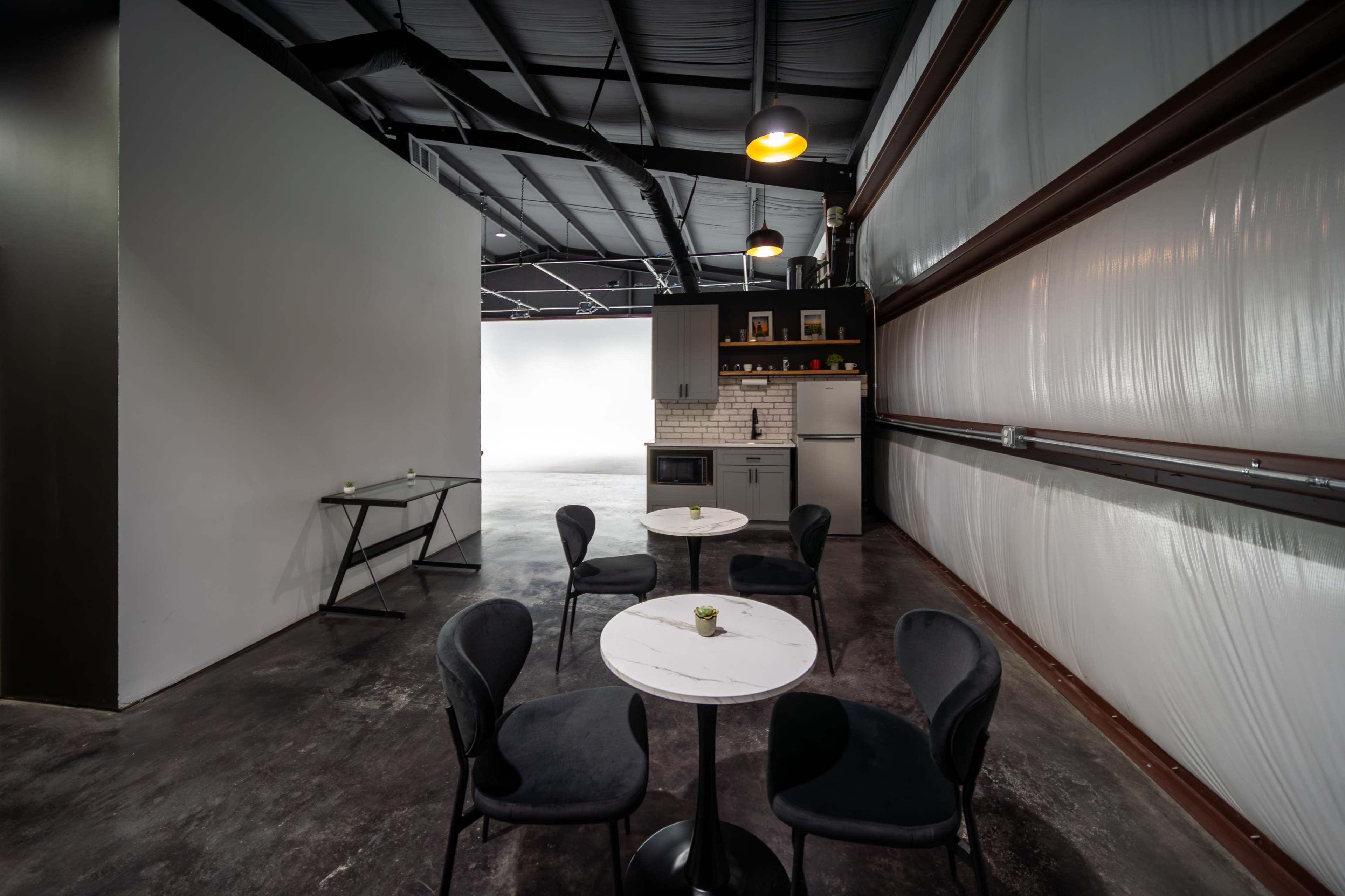 The image shows a modern interior space with two small round tables and four black chairs, accompanied by a kitchenette against a wall.