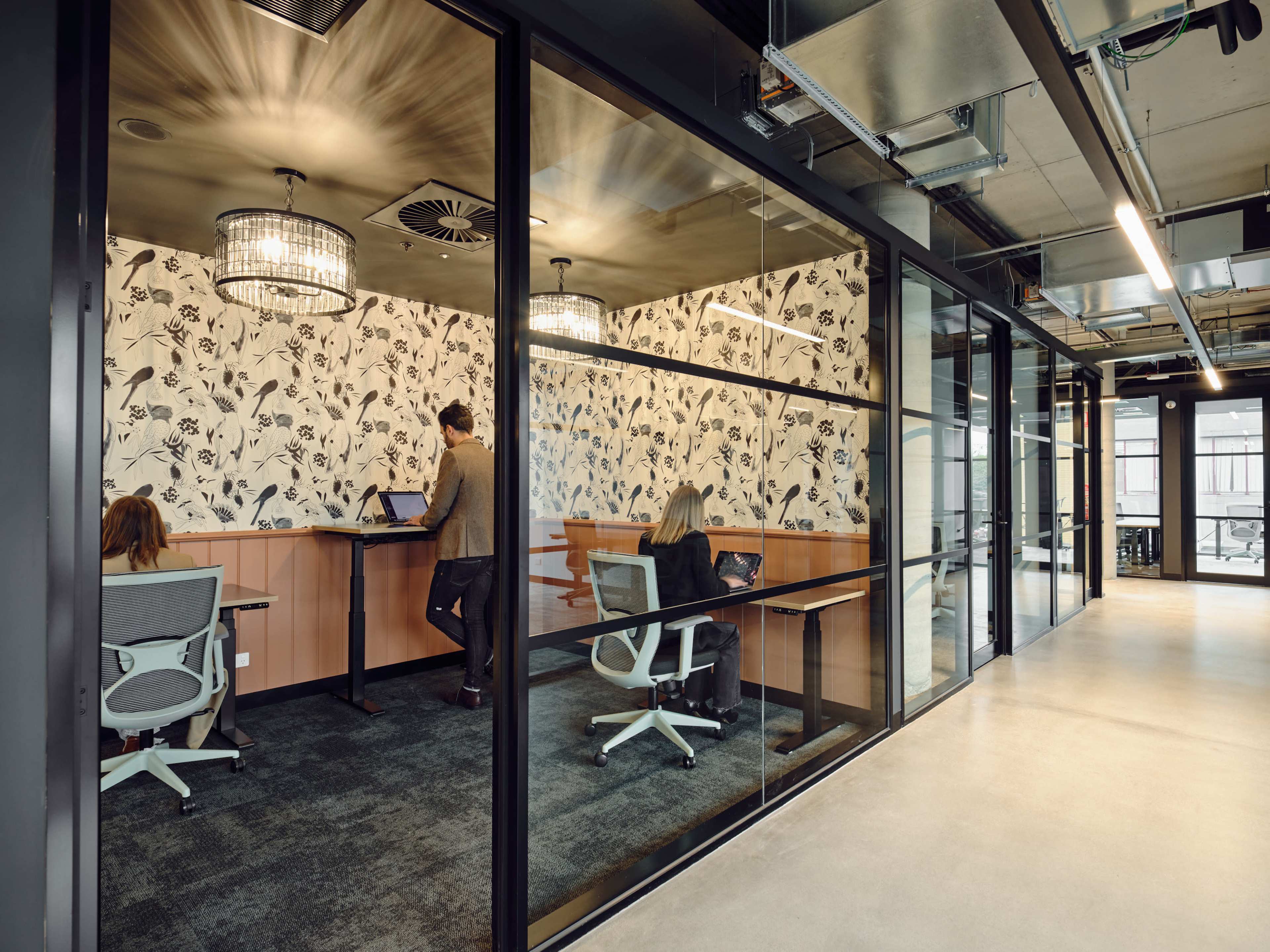 The image shows a modern office space with a glass-walled meeting area, featuring three people working at desks and floral wallpaper.