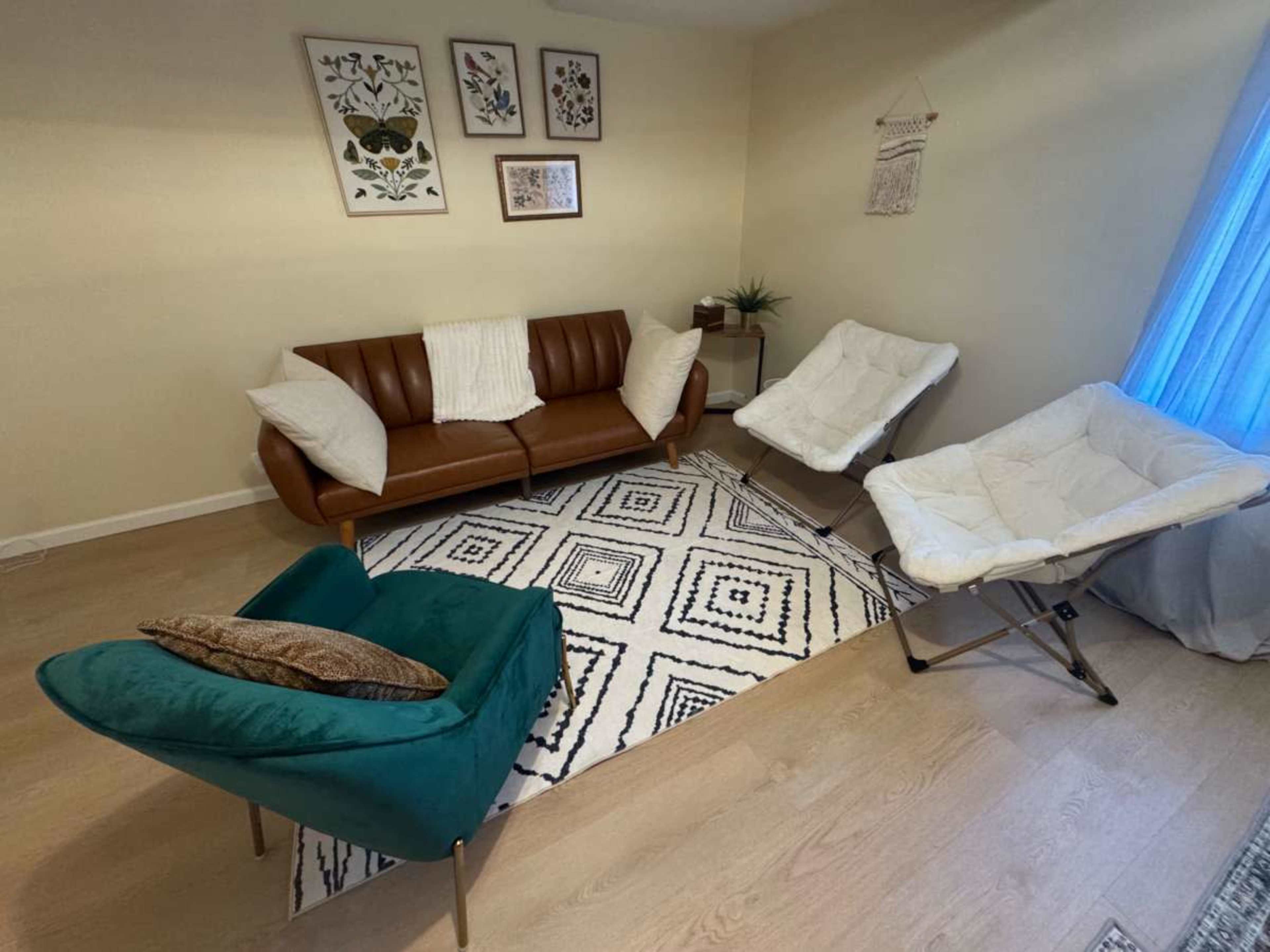 The image shows a living room with a brown leather sofa, two white folding chairs, a green accent chair, and several framed artworks on the walls.