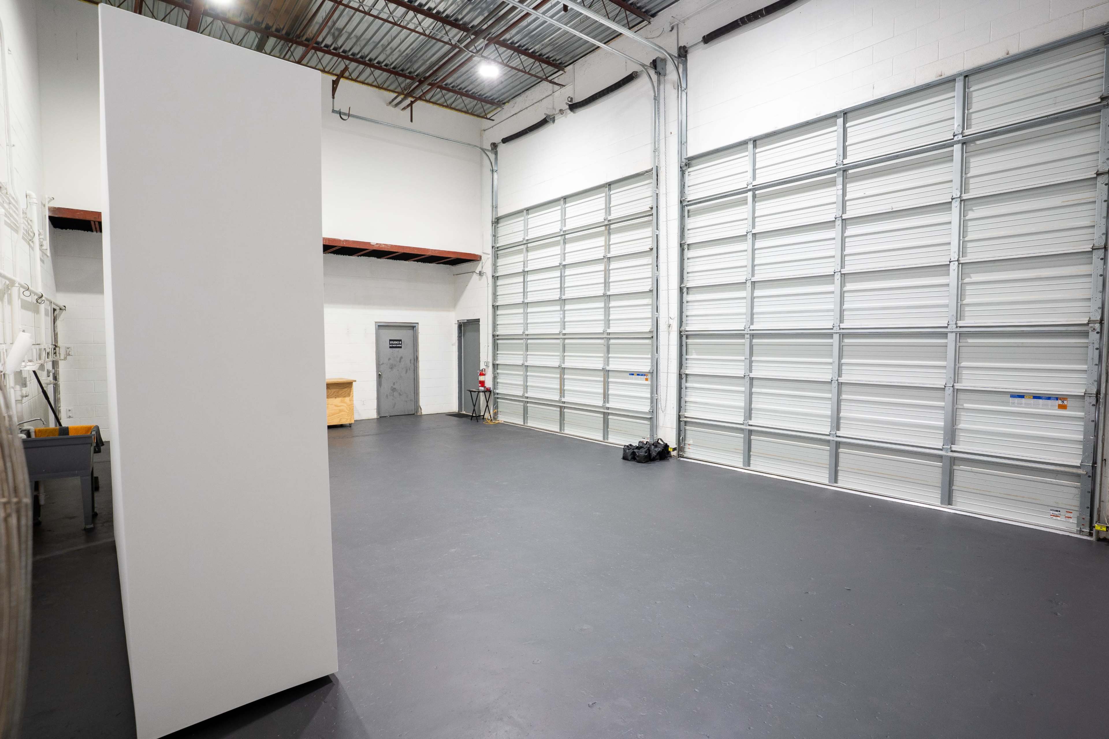 The image shows an empty industrial space with a concrete floor, a large garage door on one wall, and a plain white partition standing on the left side.