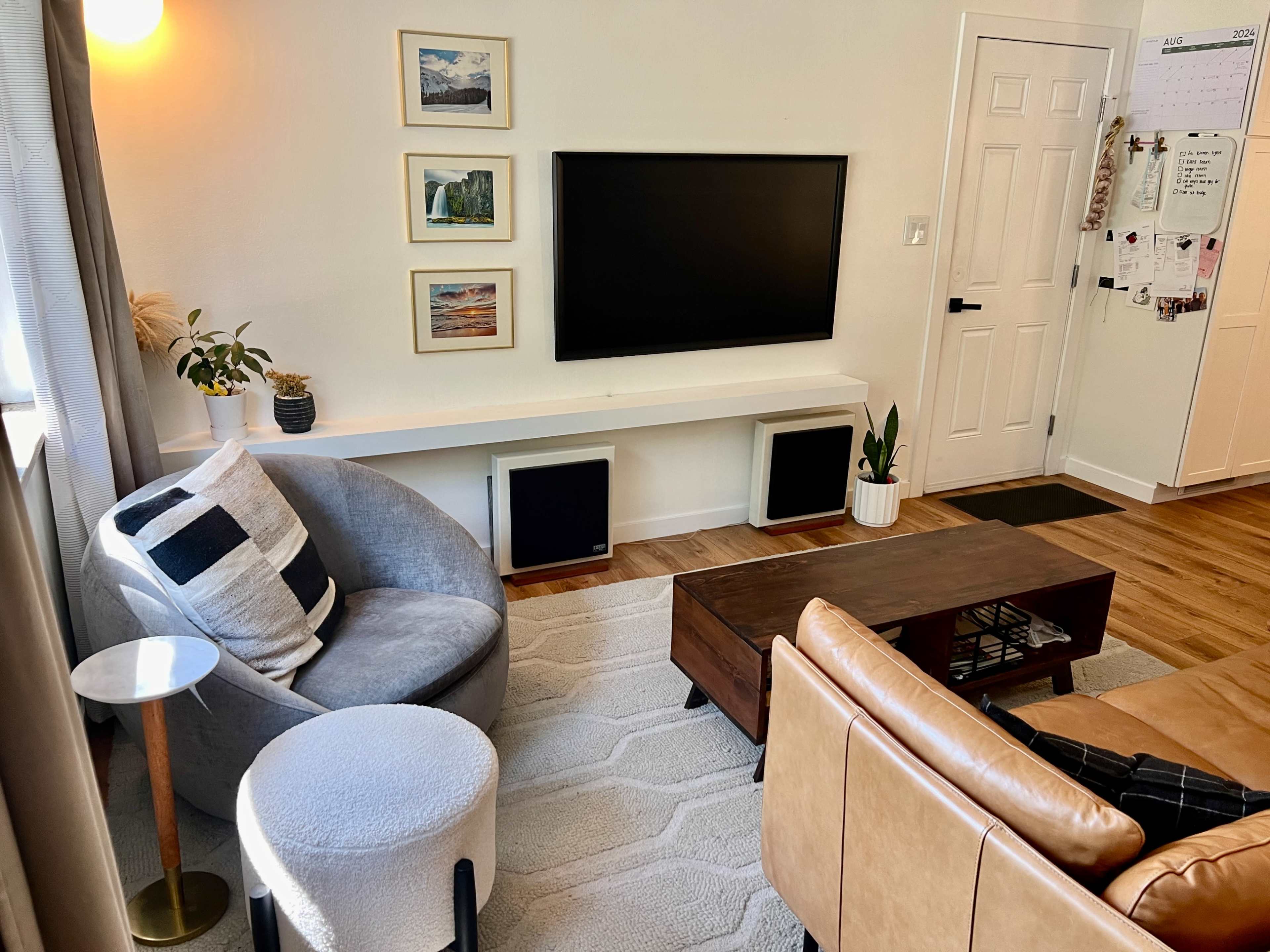 The image shows a living room with a television mounted on the wall, a coffee table, two armchairs, and a plant beside a white shelf.