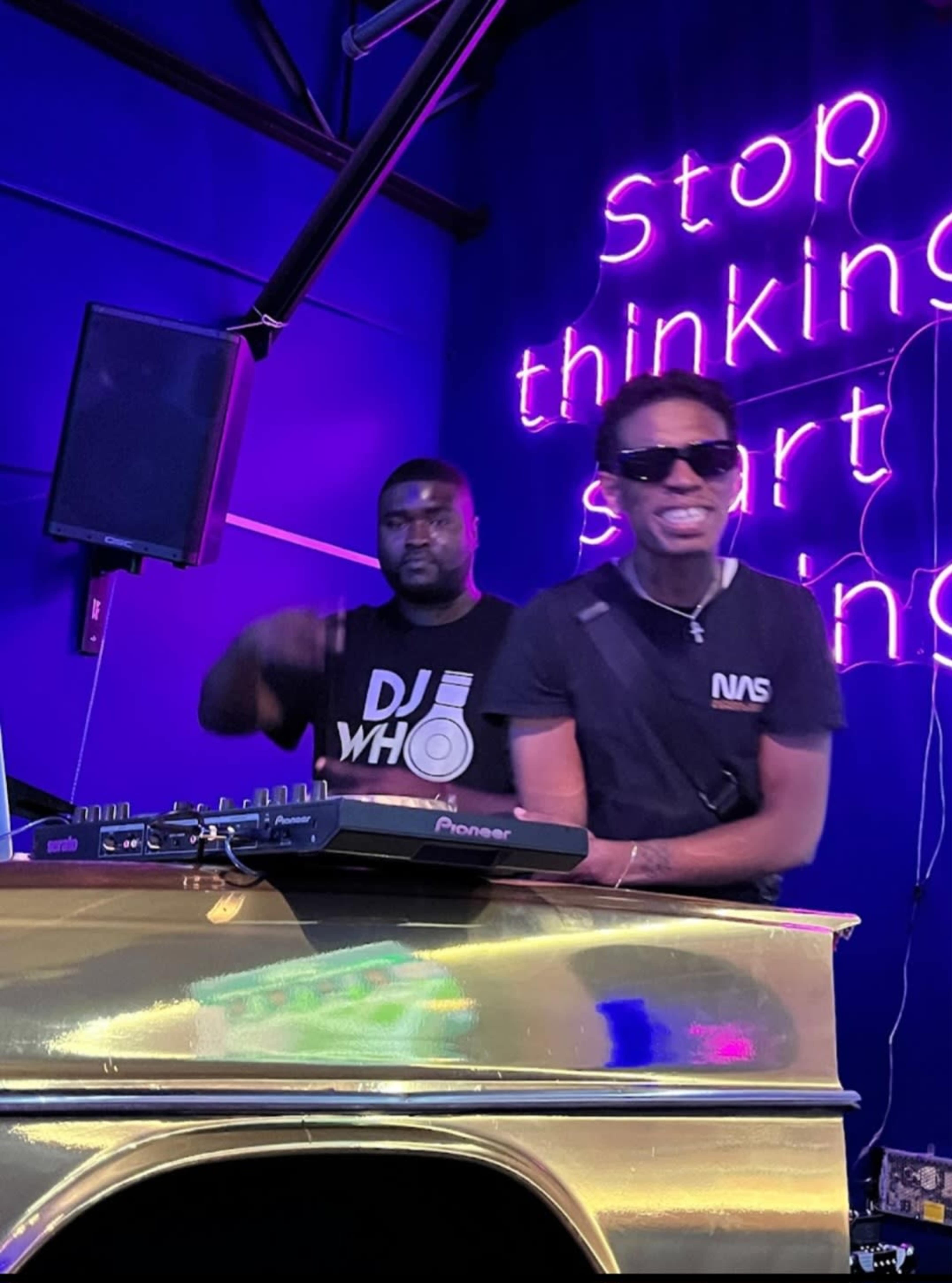 Two DJs perform on stage in front of a neon sign that reads "Stop thinking, start."