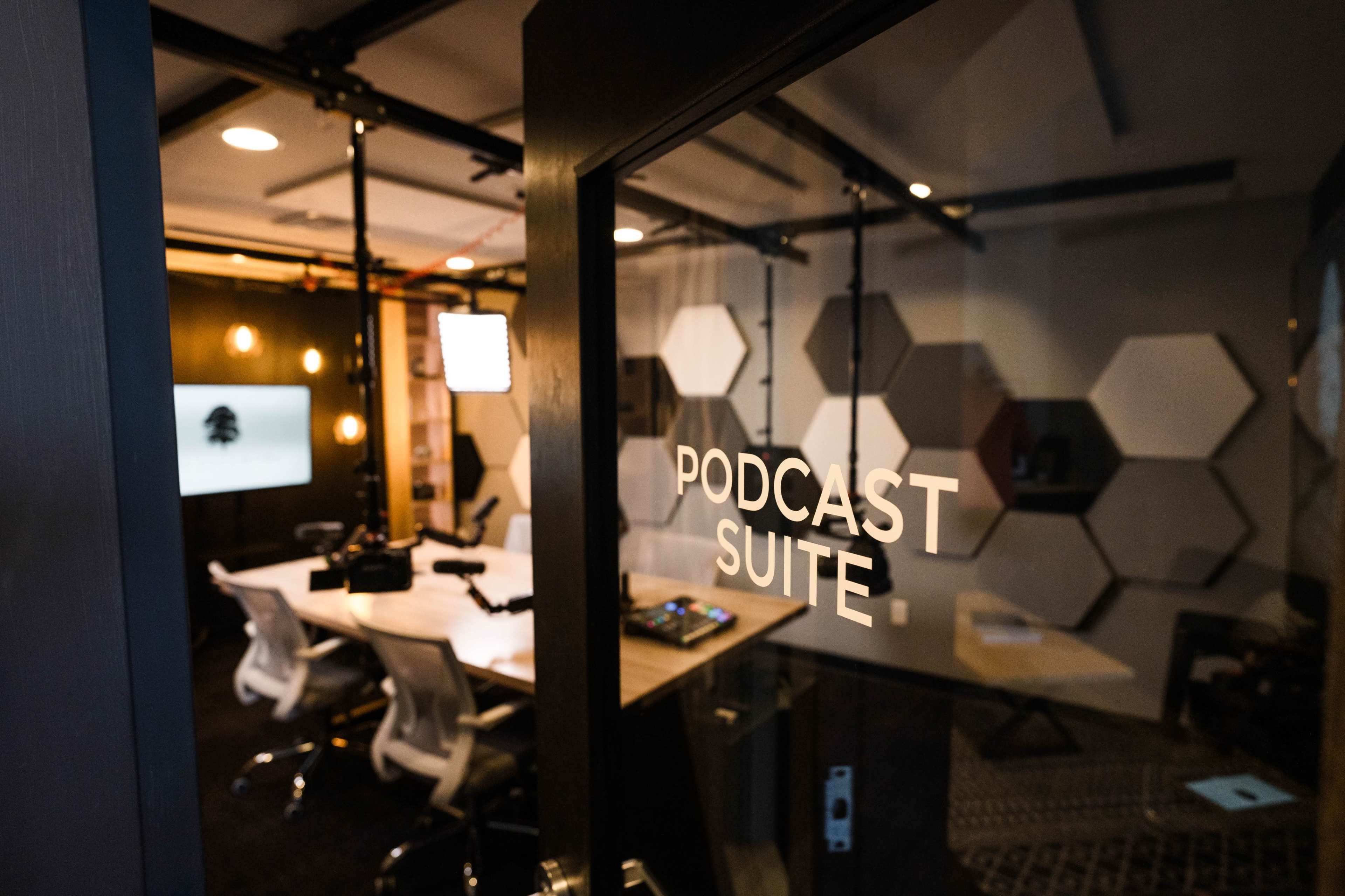 A glass door labeled "PODCAST SUITE" opens to a modern recording studio featuring a large table, chairs, and audio equipment.