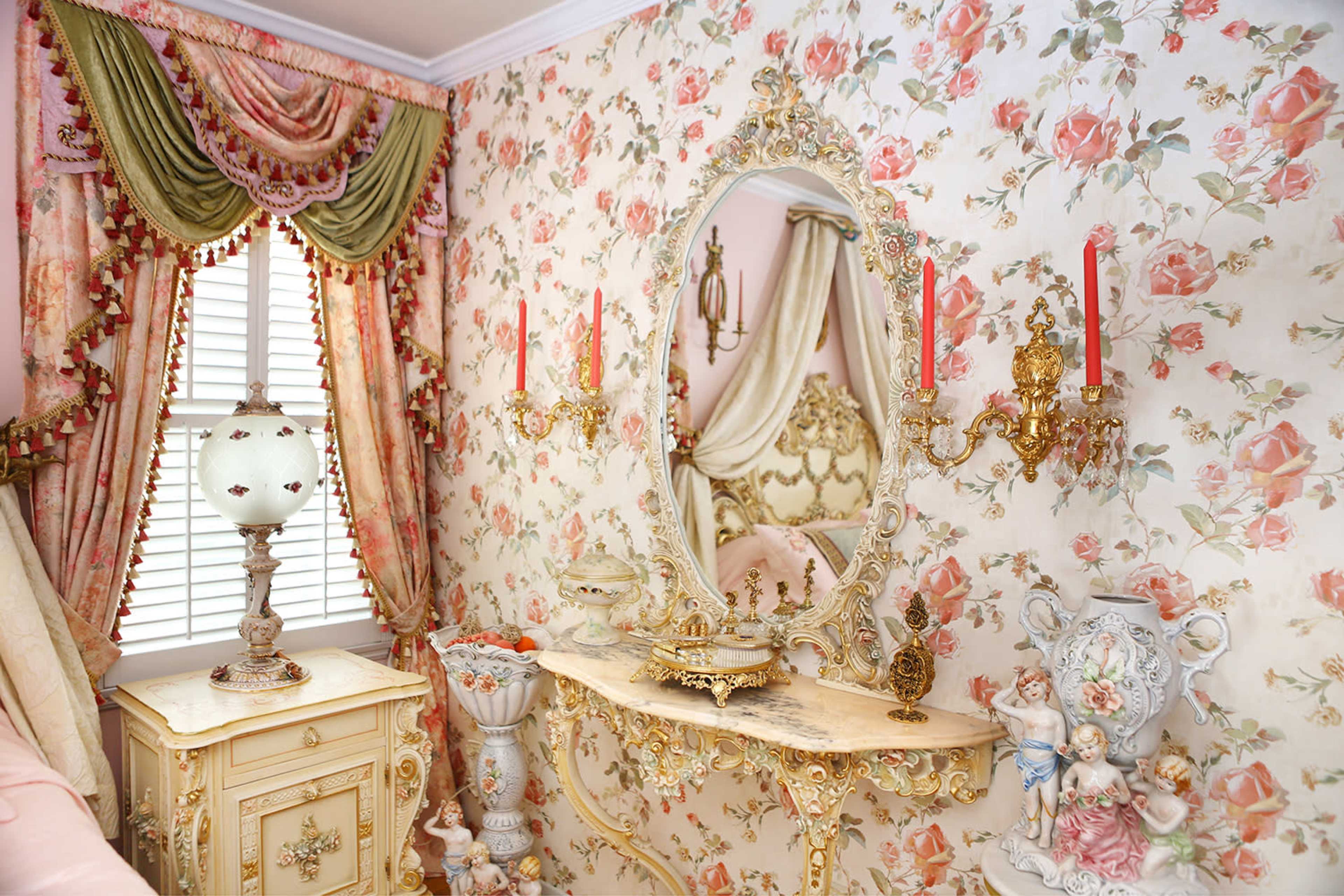 A floral-patterned room with ornate decorations, a mirror, a lamp, and candle holders against a wallpapered wall.
