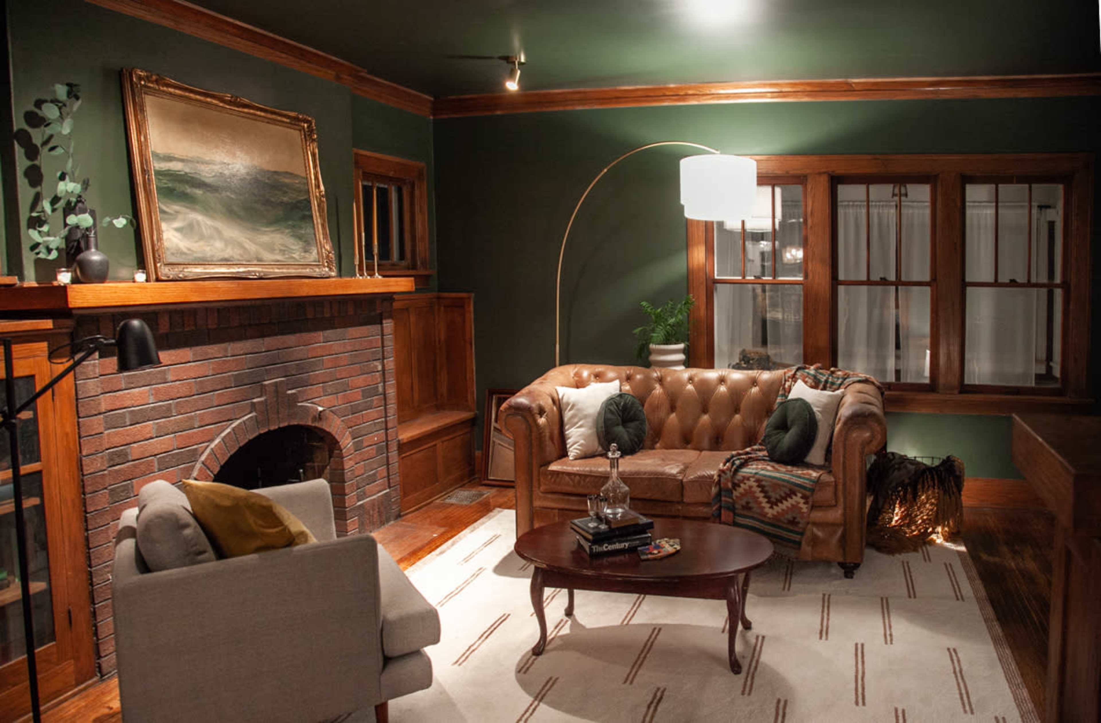A cozy living room featuring a brown leather chesterfield sofa, a round wooden coffee table, a brick fireplace, and large windows with white curtains.