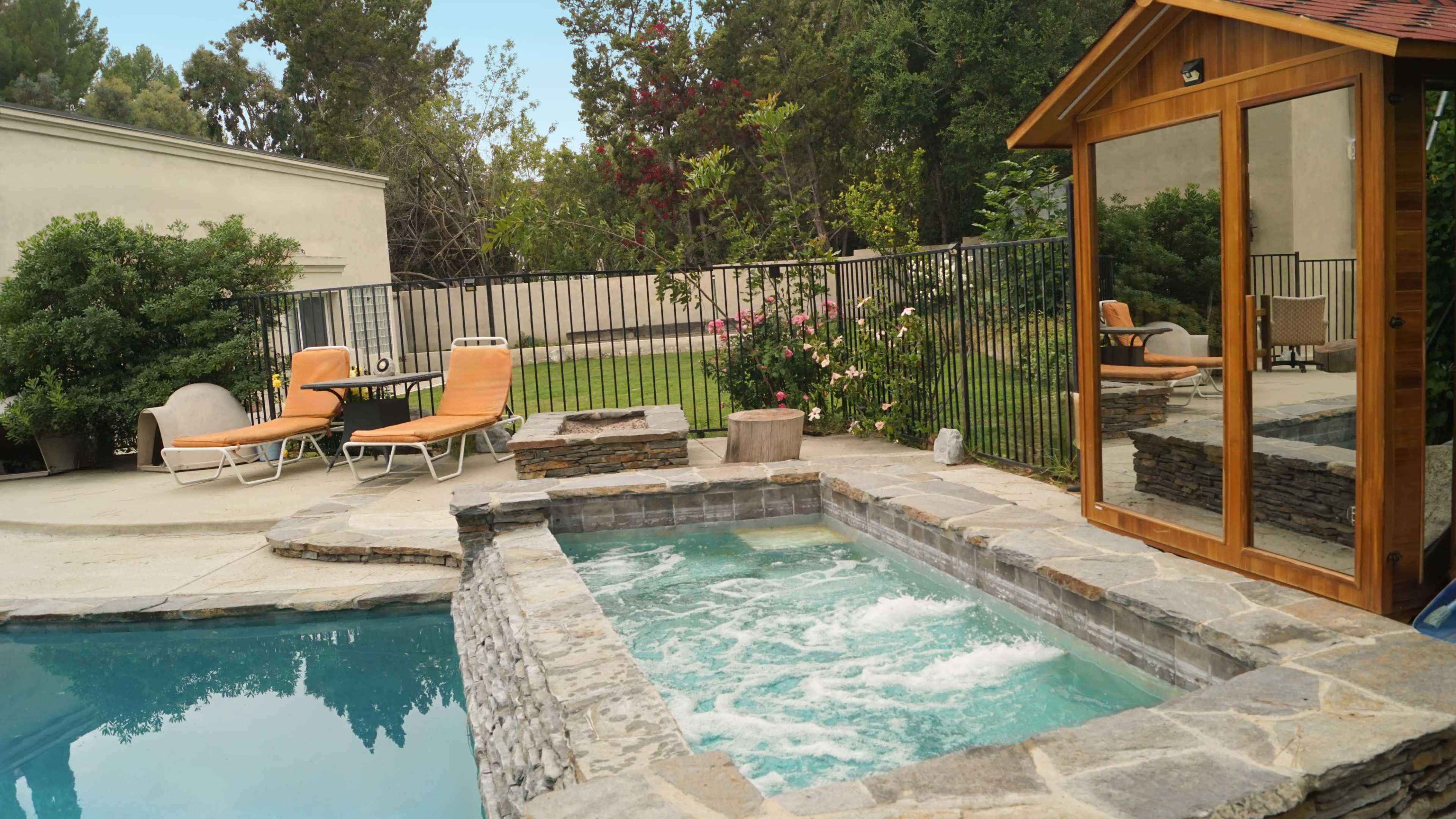 The image shows a backyard with a stone hot tub adjacent to a swimming pool, surrounded by lounge chairs and a wooden structure with a mirror.