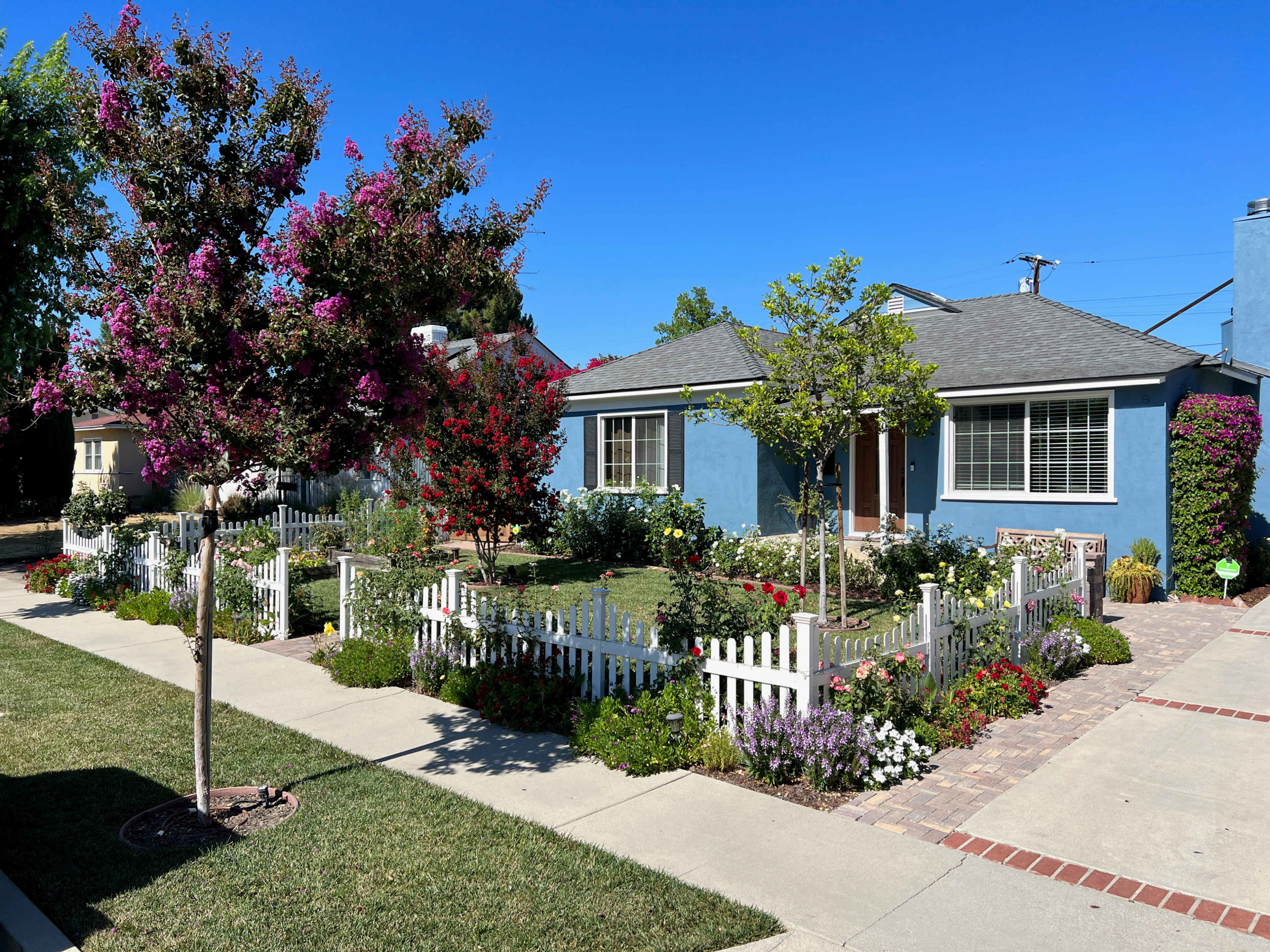 A light blue house with a gray roof is surrounded by colorful flower gardens and a white picket fence.