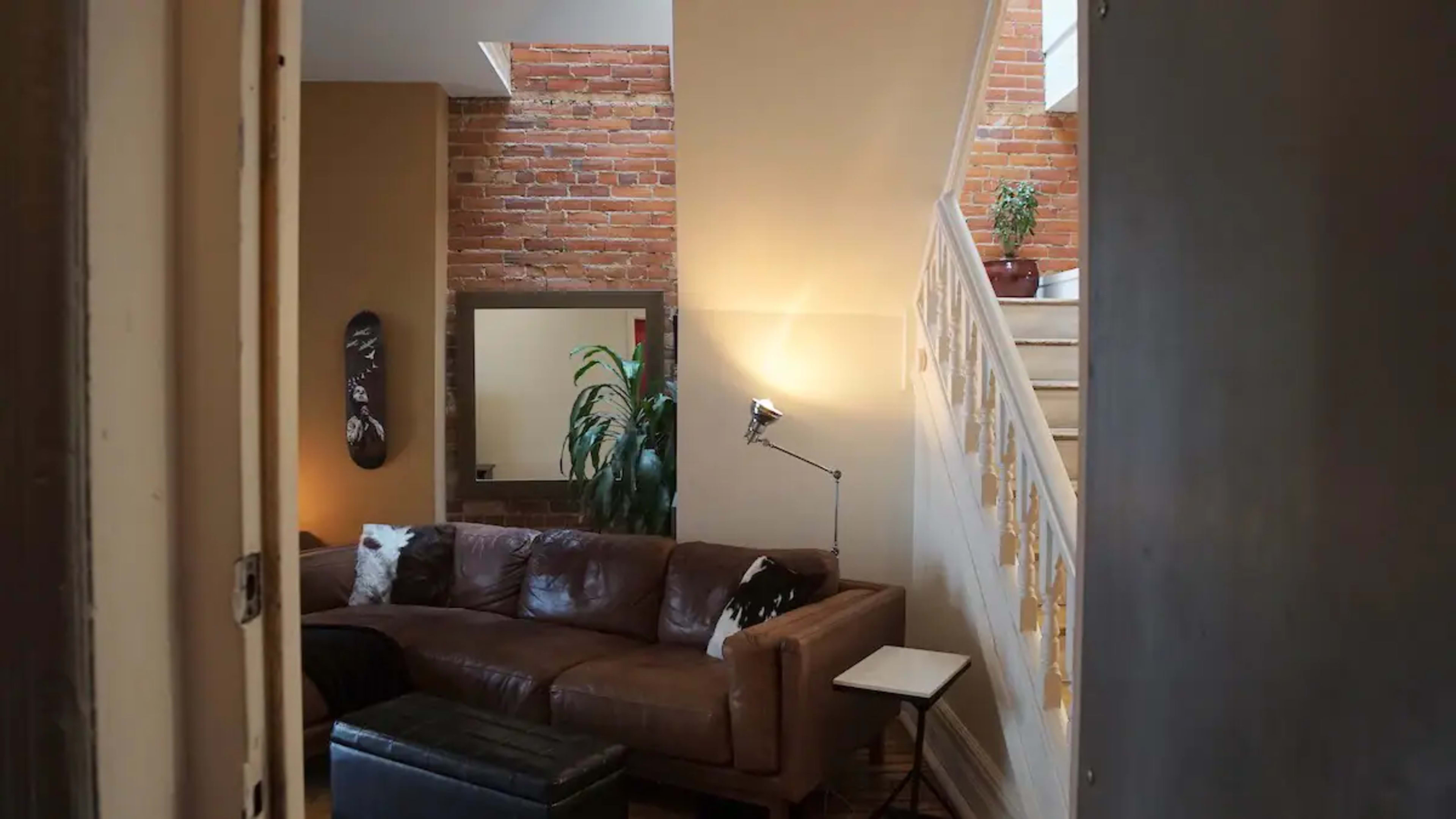 The image shows a cozy living room with a brown leather sectional sofa, a staircase leading to an upper level, and a large mirror on the wall next to a potted plant.