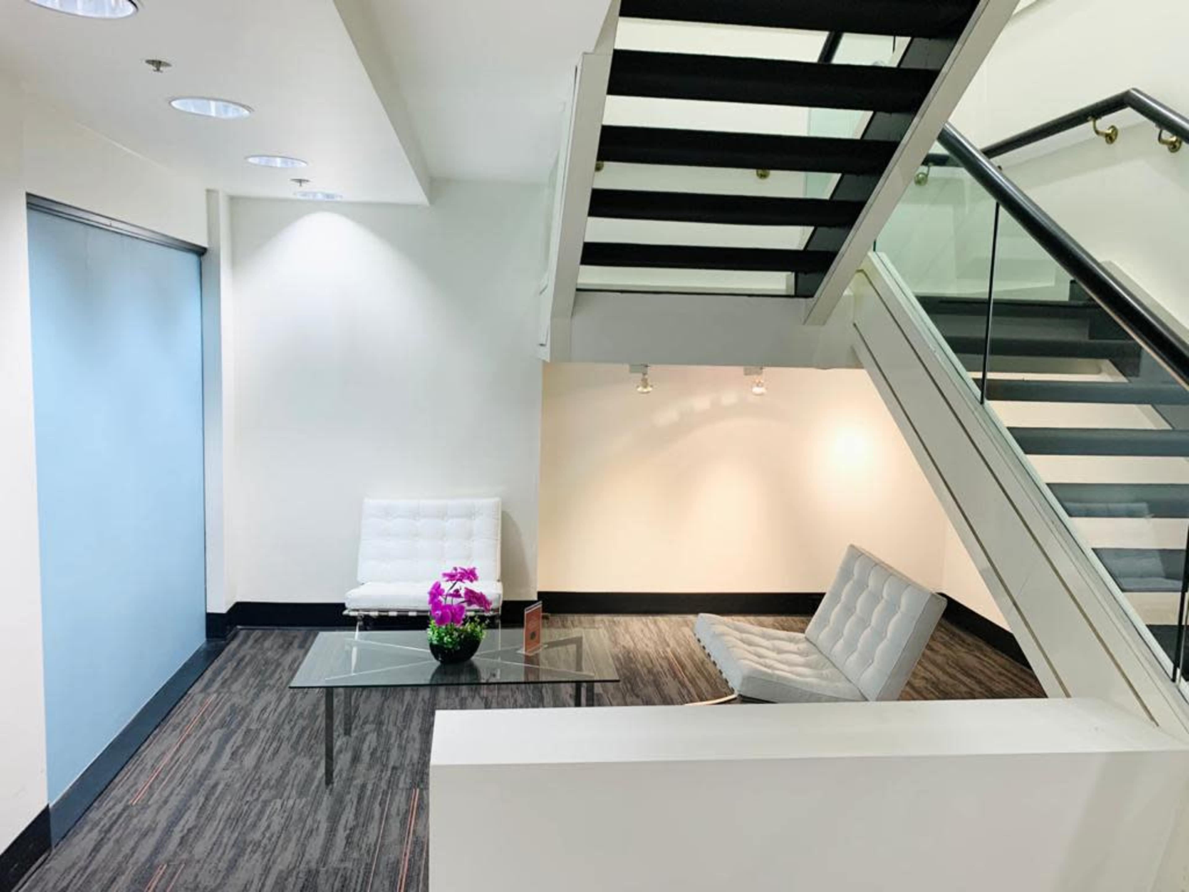 A modern, minimalistic lobby area featuring a glass table, two white chairs, and a staircase leading to a second level.