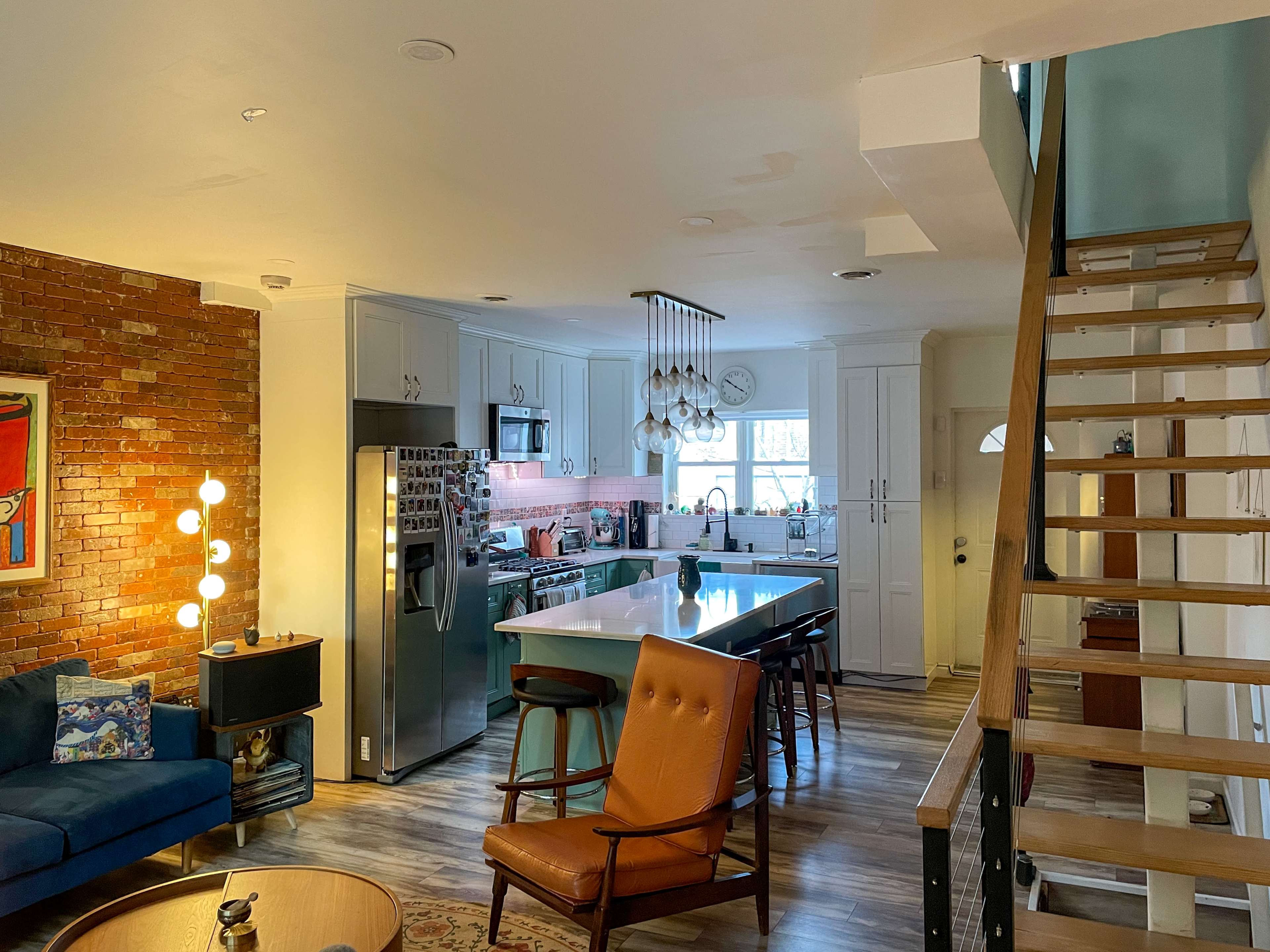 The image shows a modern open-concept kitchen and living area with a staircase on the right and a brick accent wall.