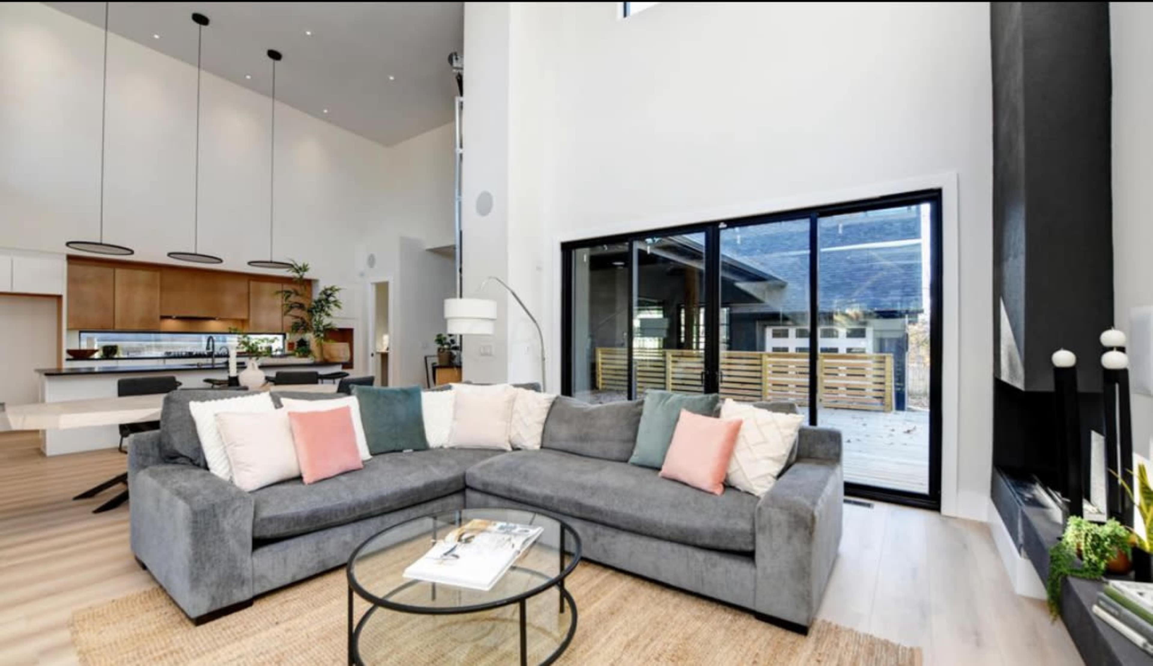 A modern living room featuring a gray sectional sofa with colorful pillows, a glass coffee table, and large windows that allow sunlight to enter the space.
