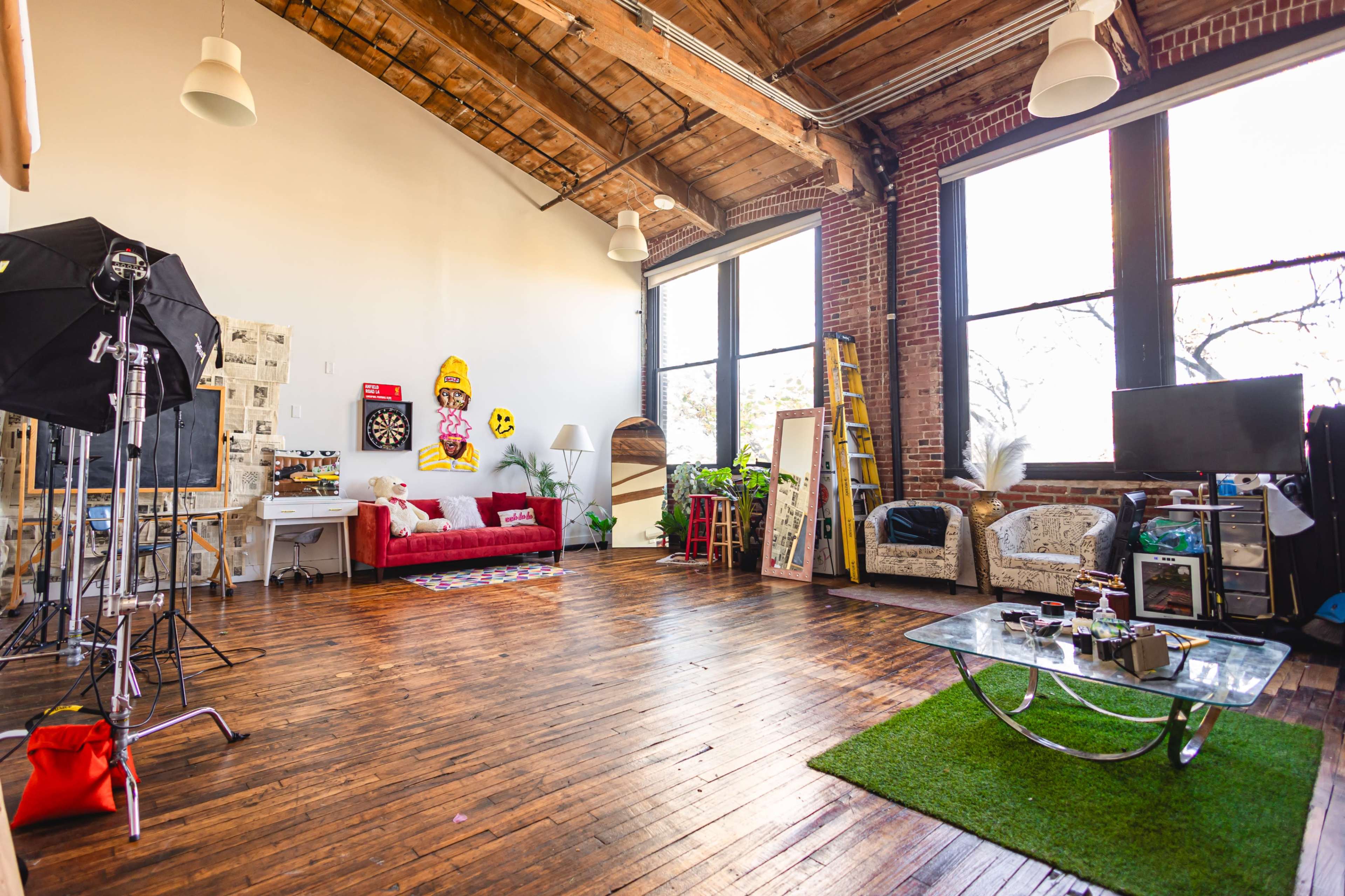 The image shows an open loft space with wooden floors, large windows, a red couch, a dartboard on the wall, and various furniture and decorations scattered throughout.