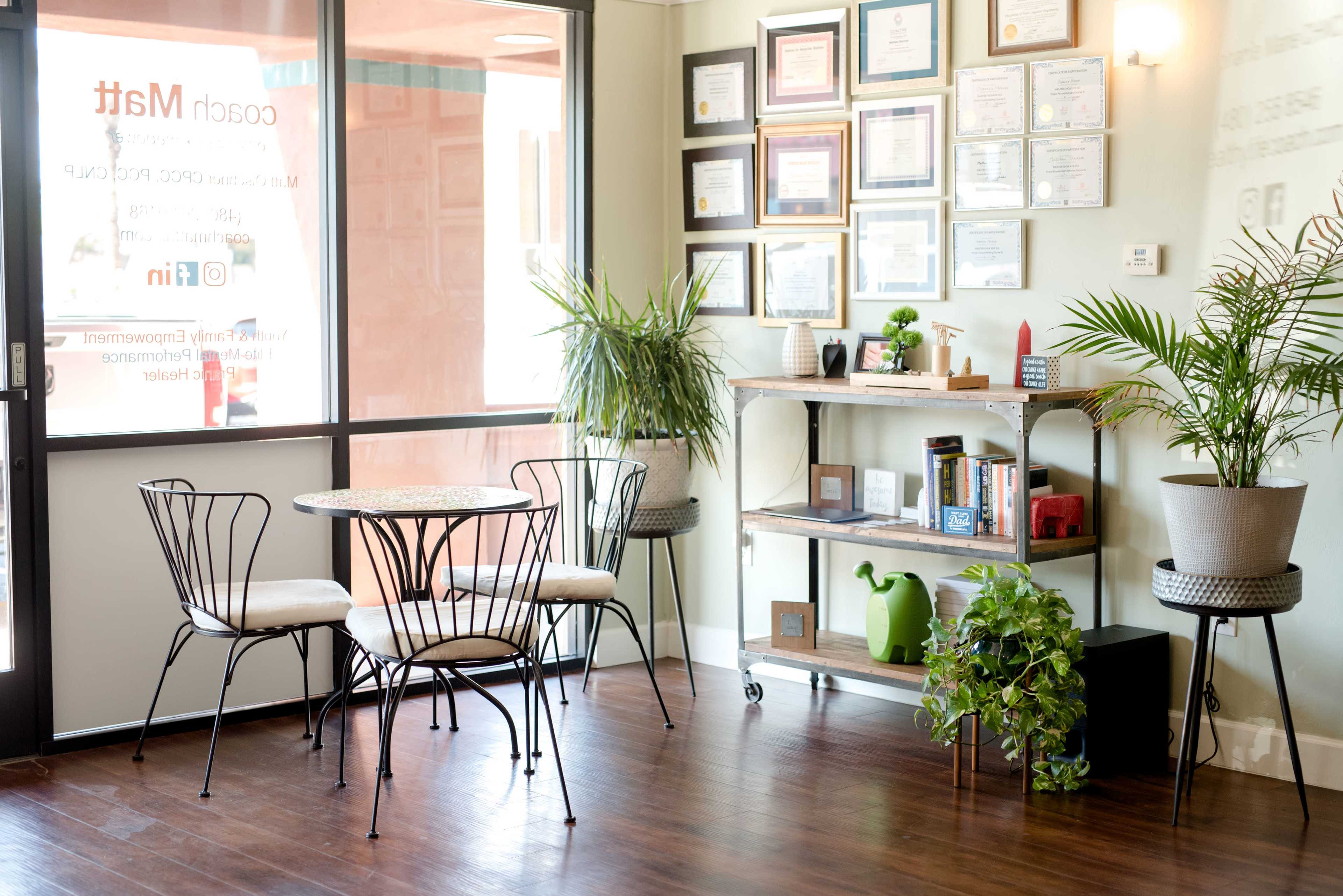 A small waiting area features a round table with four chairs, a plant stand, and a wall displaying various framed certificates.