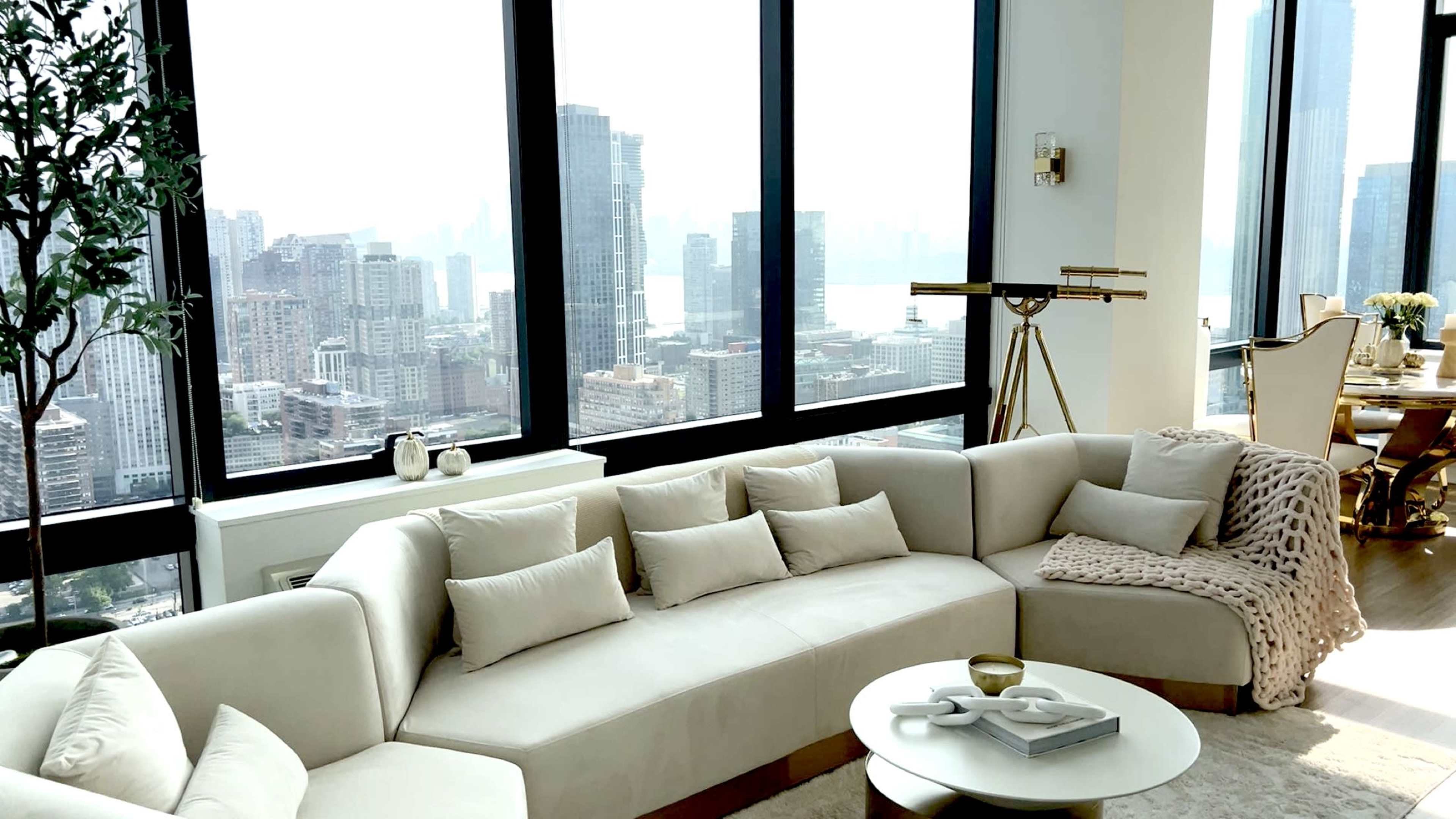 The image shows a modern living room with a curved beige sofa, numerous pillows, a round coffee table, and large windows overlooking a city skyline.