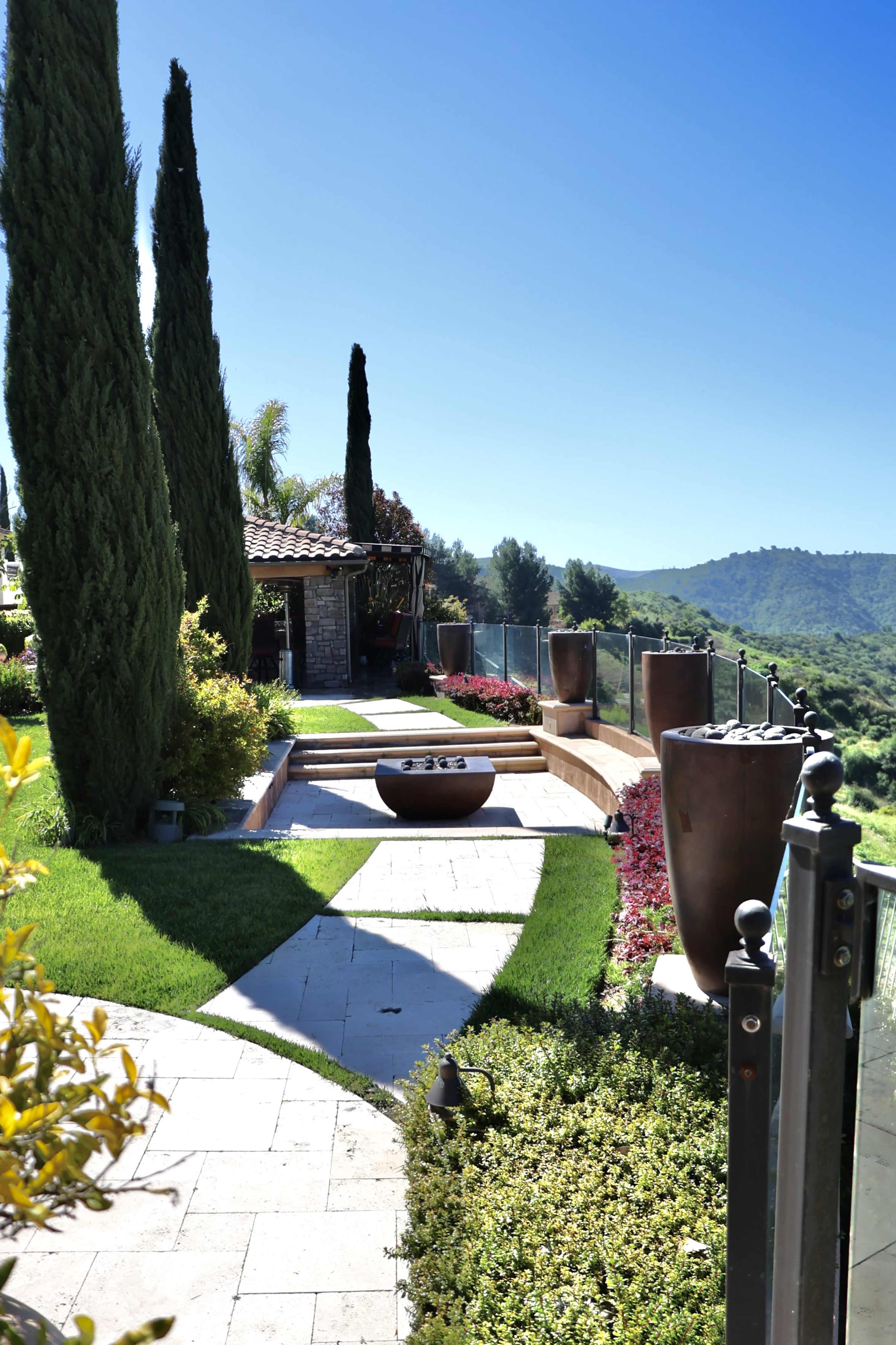 A landscaped garden featuring stone pathways, large planters, and a fire pit, set against a backdrop of rolling hills under a clear blue sky.