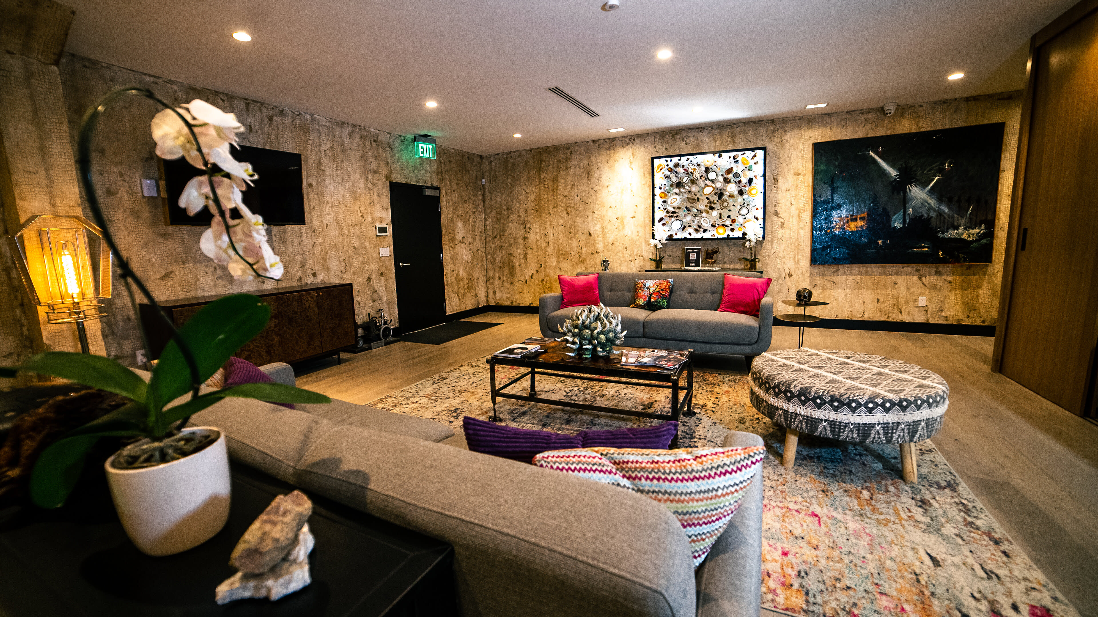 A modern living room with a gray sofa, decorative pillows, a round patterned ottoman, an orchid plant, and artwork on the walls.