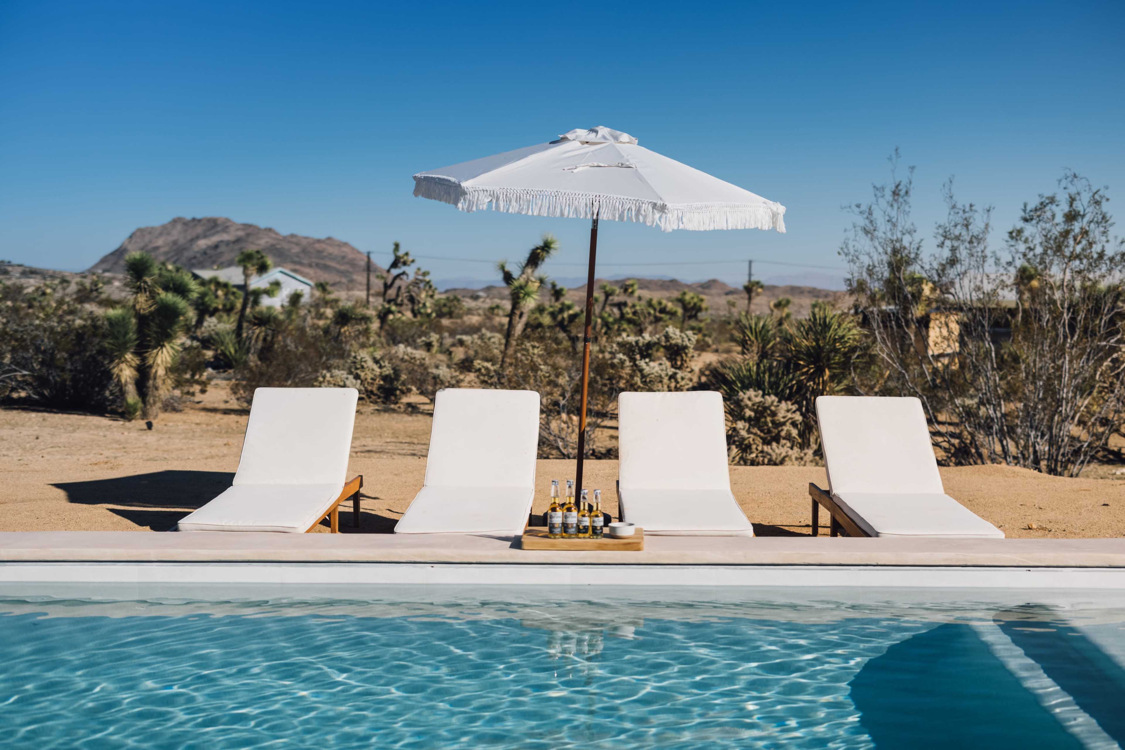 Four lounge chairs beside a swimming pool, shaded by a white umbrella, with a desert landscape and mountains in the background.