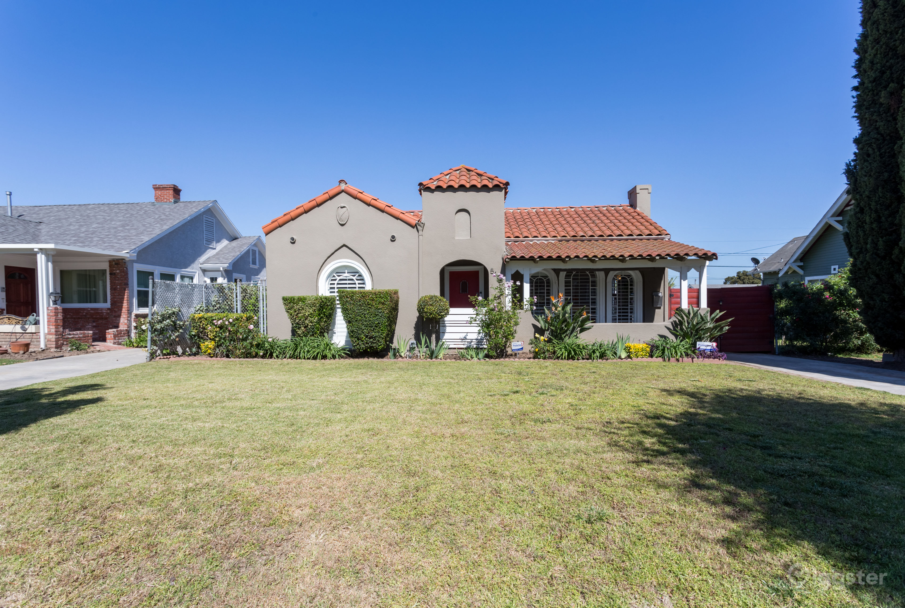 The image shows a single-story house with a red tile roof, a rounded front arch, and well-maintained landscaping on a clear day.