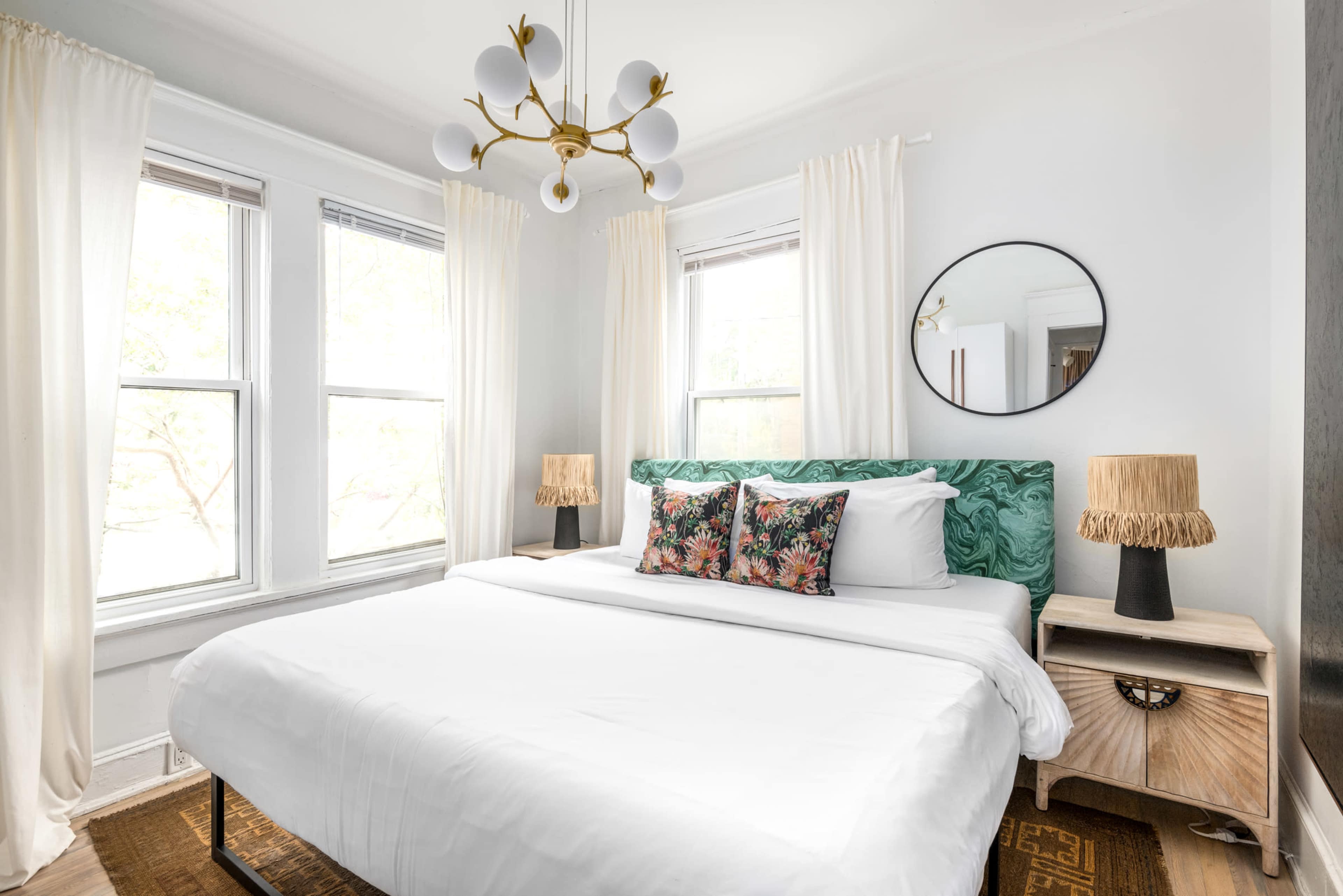 The image shows a bright bedroom with a large bed featuring a patterned green headboard, two bedside lamps, and large windows with sheer curtains.