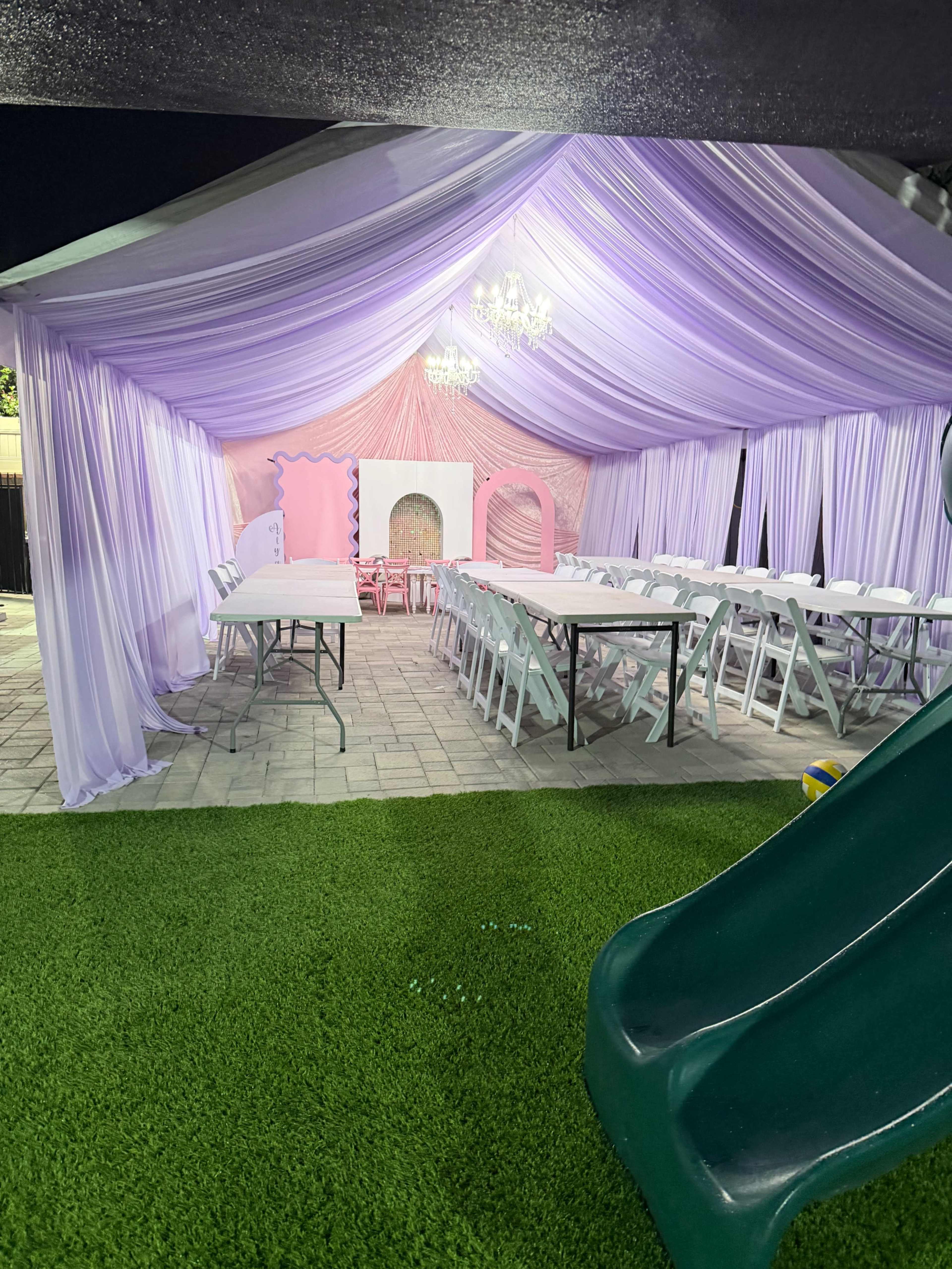 A decorated outdoor tent features folding tables and chairs, draped fabric in lavender and pink, and a grassy area with a slide in the foreground.