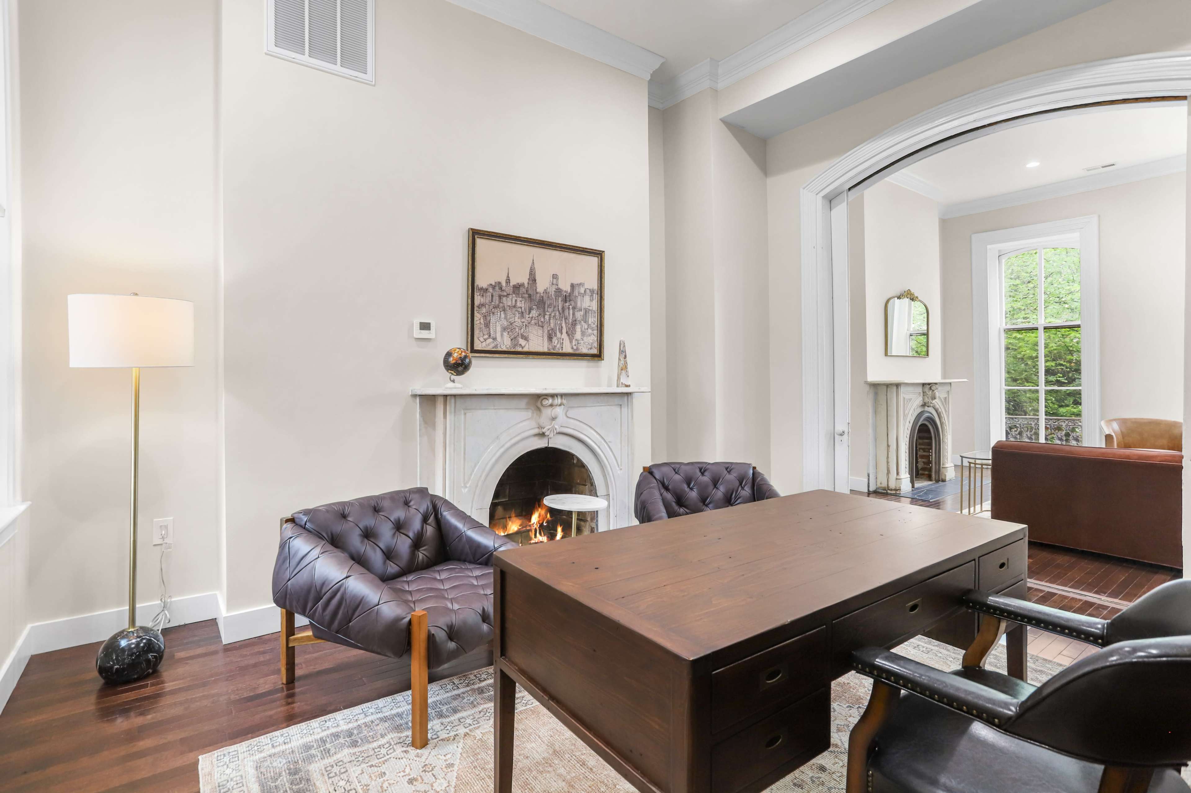 The image shows a well-lit room featuring a plush brown armchair, a wooden desk, and a fireplace with a small flame, framed by large windows.