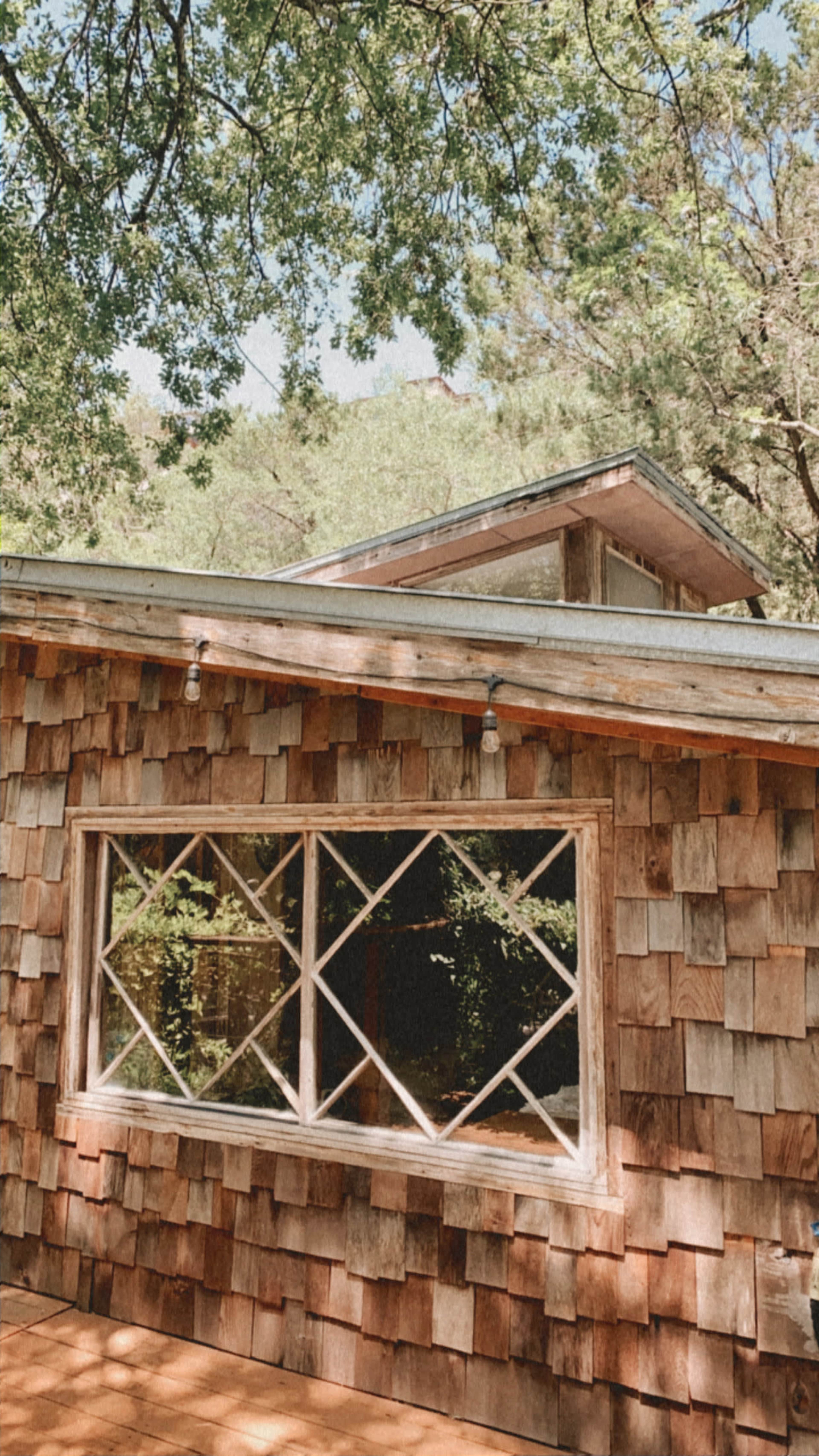The image shows a wooden cabin with a shingled exterior and a large window featuring a diamond-pane design, surrounded by trees.