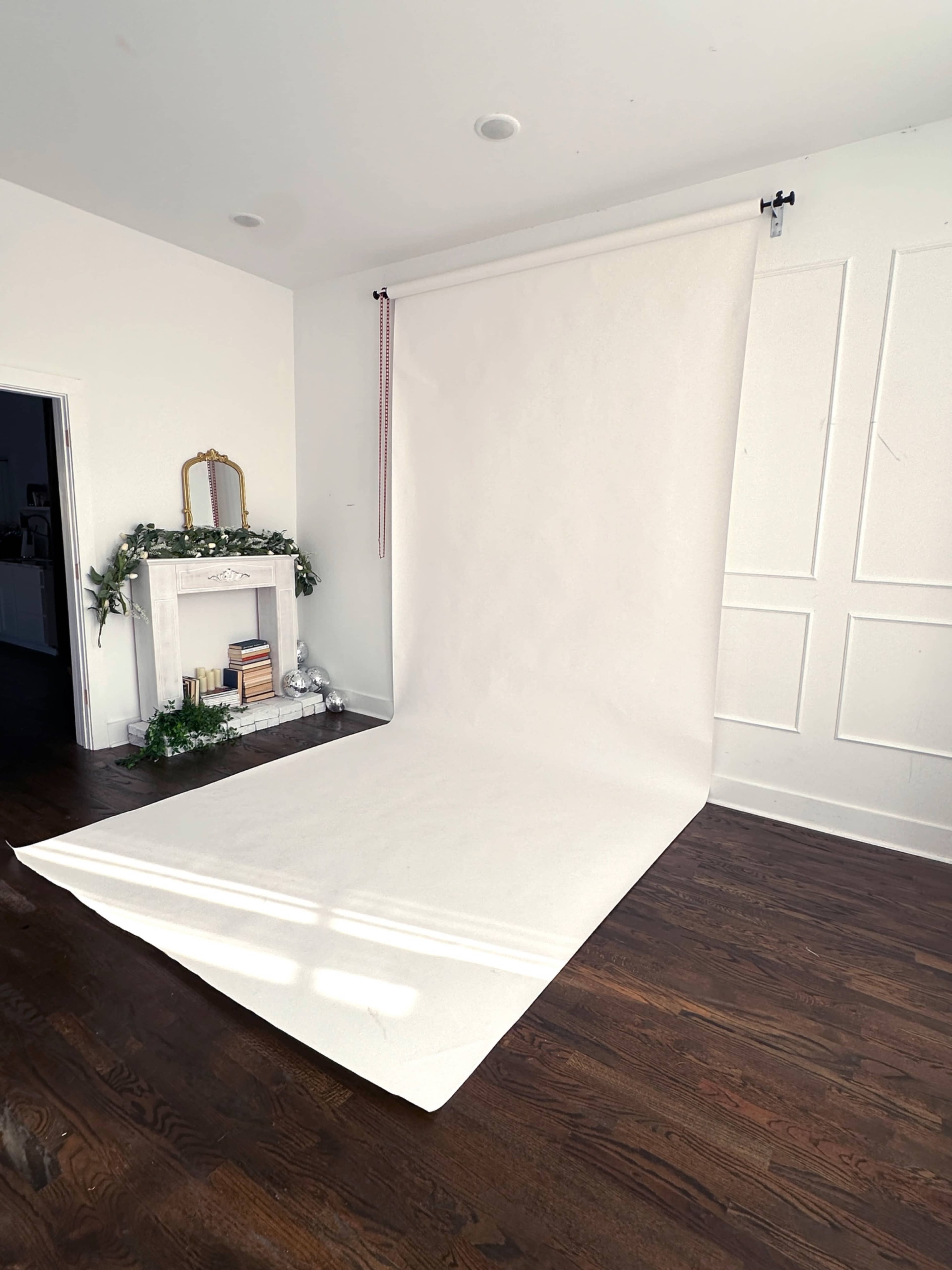 The image shows a spacious room with a white backdrop set up against a wall, wooden flooring, and a decorative fireplace.