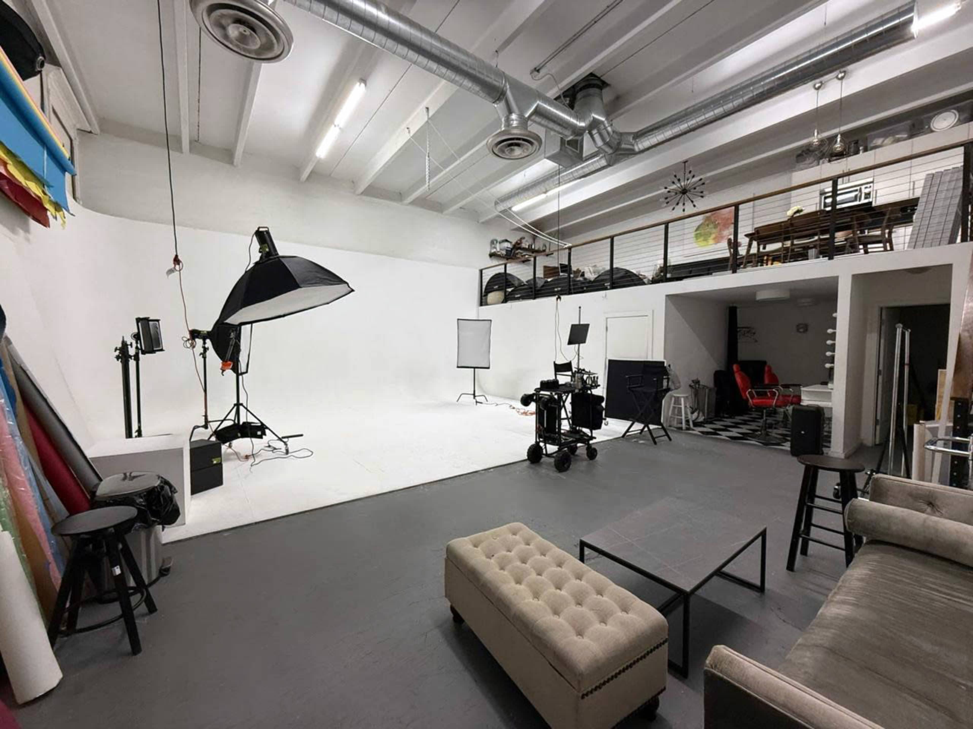 The image shows a spacious photography studio with a white backdrop, lighting equipment, and various furniture arranged around the room.