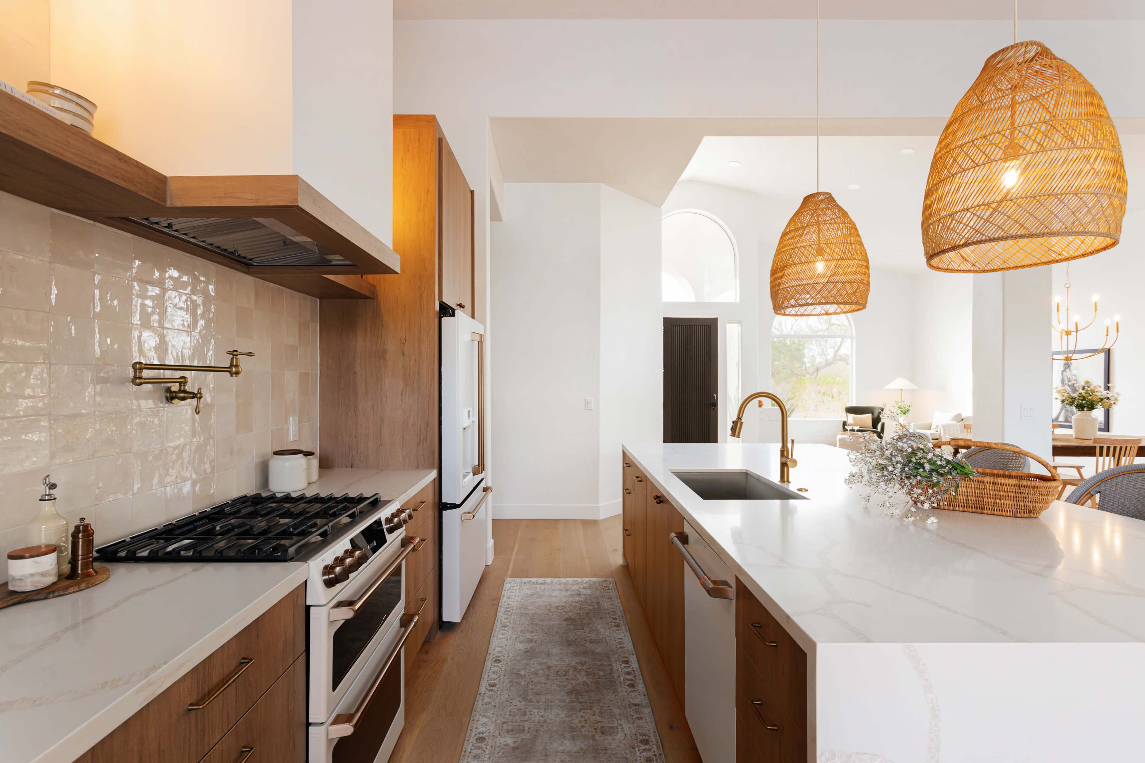 The image shows a modern kitchen with wooden cabinets, a large island featuring a sink, and pendant lights hanging above.