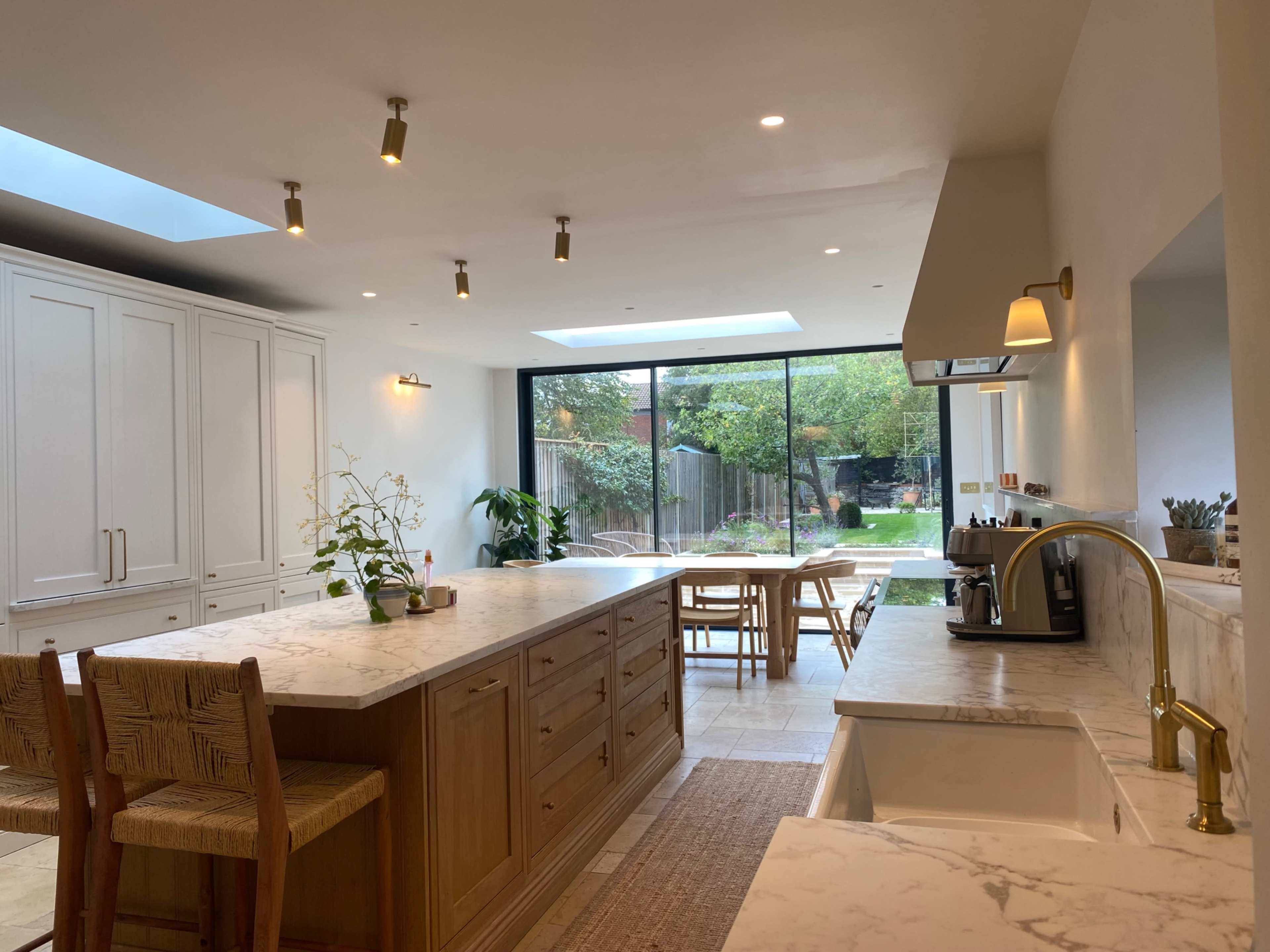 The image shows a modern kitchen with a large island, marble countertops, and a view of a garden through a glass wall.