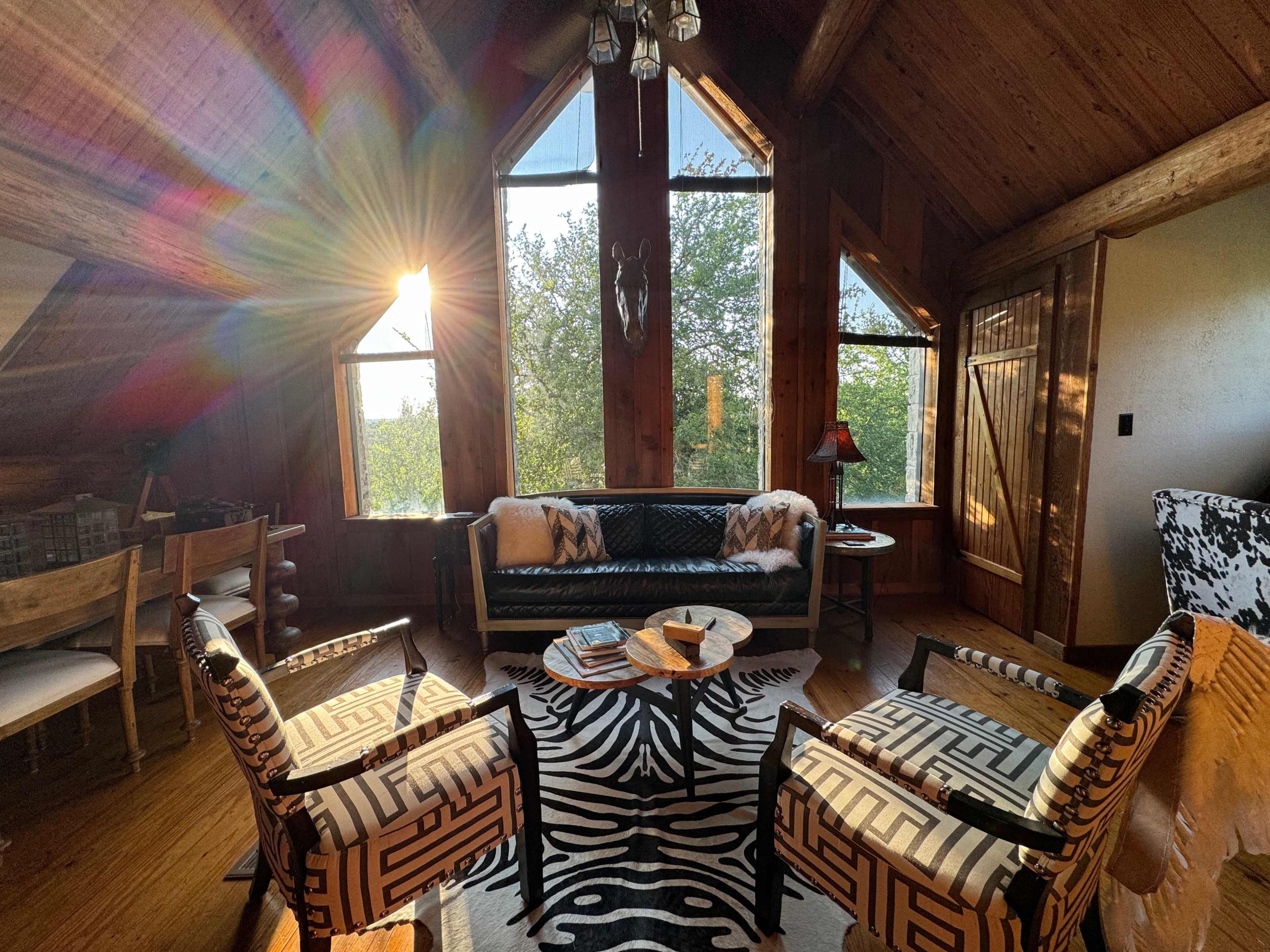A sunlit living room features a black leather sofa, two patterned chairs, and large windows overlooking a natural landscape.